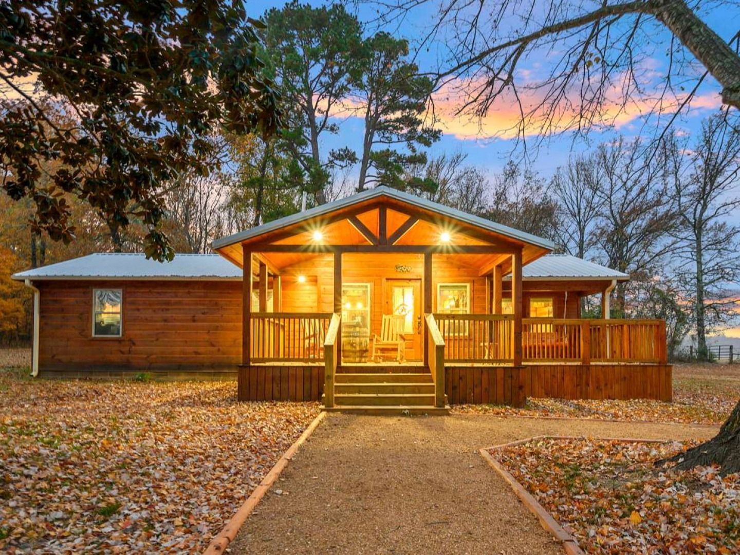 Cozy lit cabin at dusk with a covered porch, wooden walkway, and fallen leaves in the yard