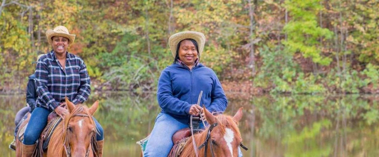 Two riders on horses in blue jackets, smiling on a wooded trail in autumn