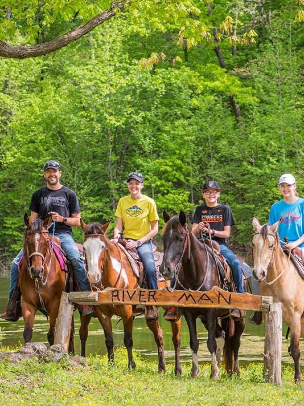 Four riders on horses posing behind a wooden River Ridge sign in a green wooded area