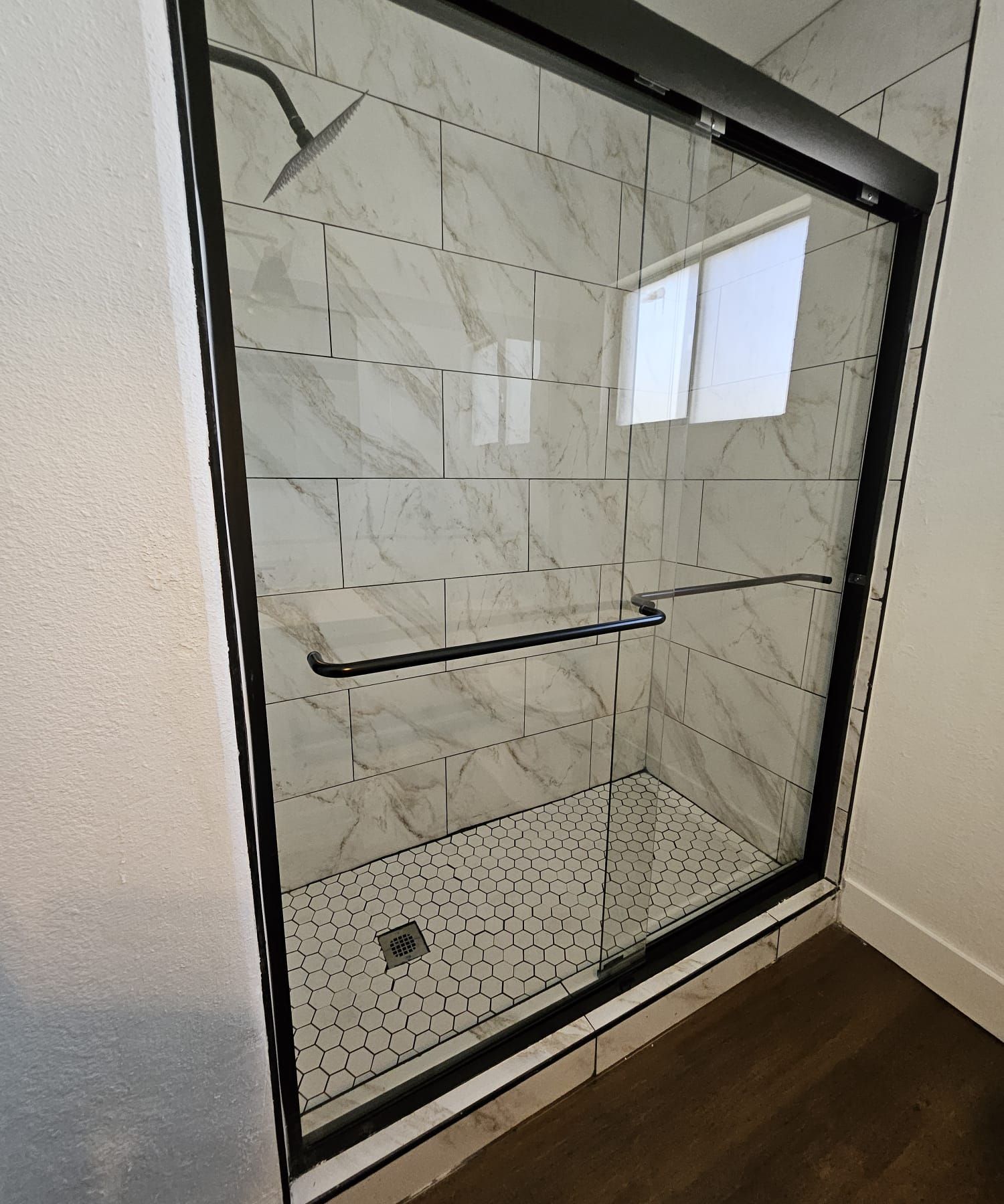 Modern shower enclosure with marble-look tile, black trim, grab bar, and small window