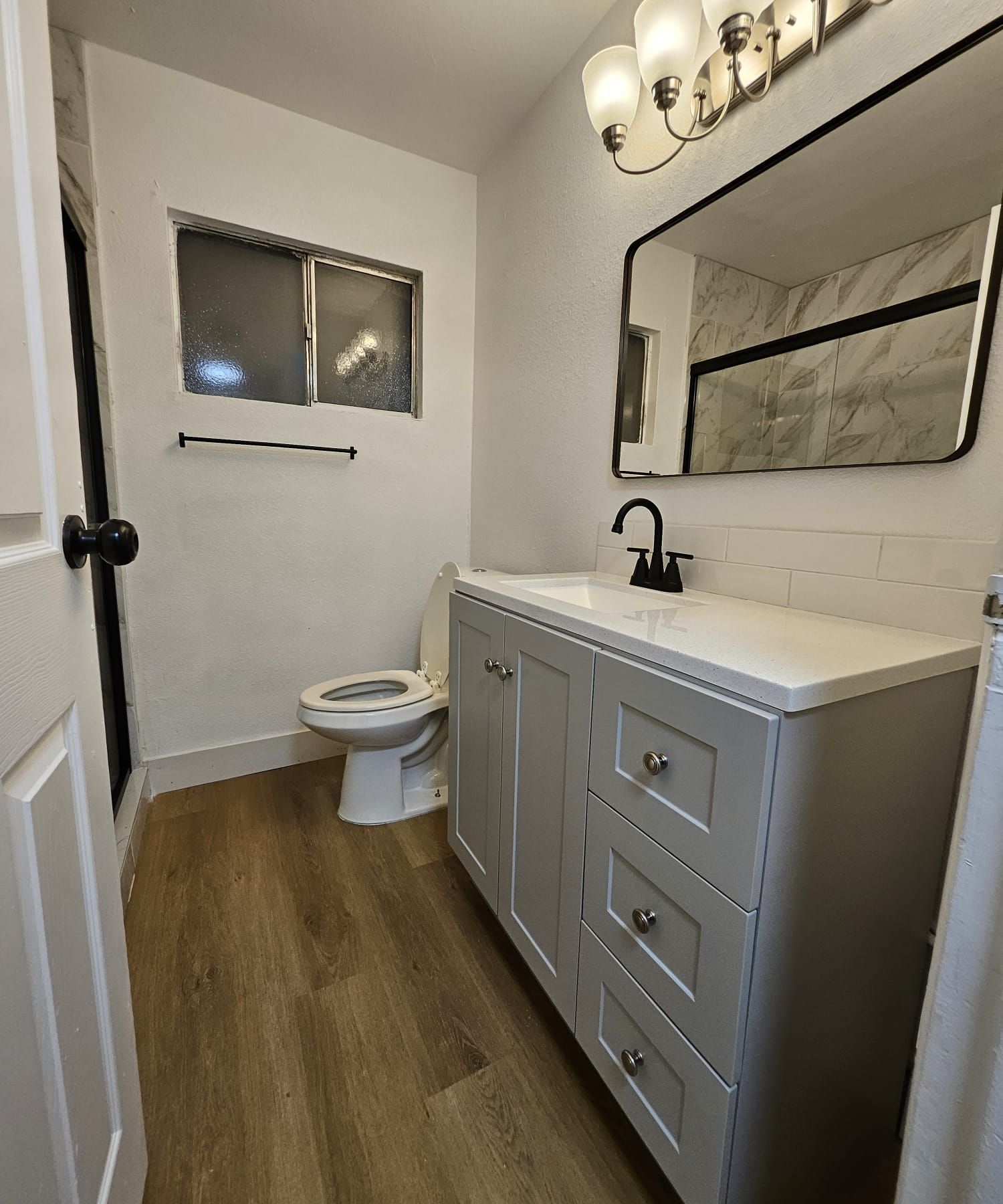 Small modern bathroom with gray vanity, black faucet, mirror, toilet, window, and wood-look floor.