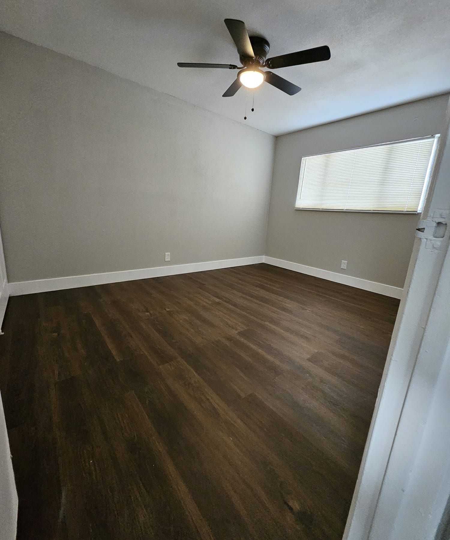 Empty bedroom with dark wood floors, gray walls, a ceiling fan, and a window with blinds.