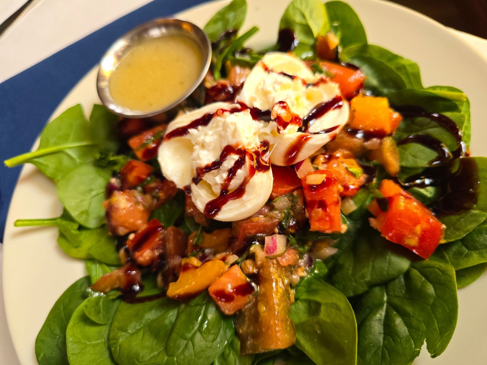 A spinach salad topped with fresh mozzarella, chopped tomatoes, and balsamic glaze, served with a side of dressing.