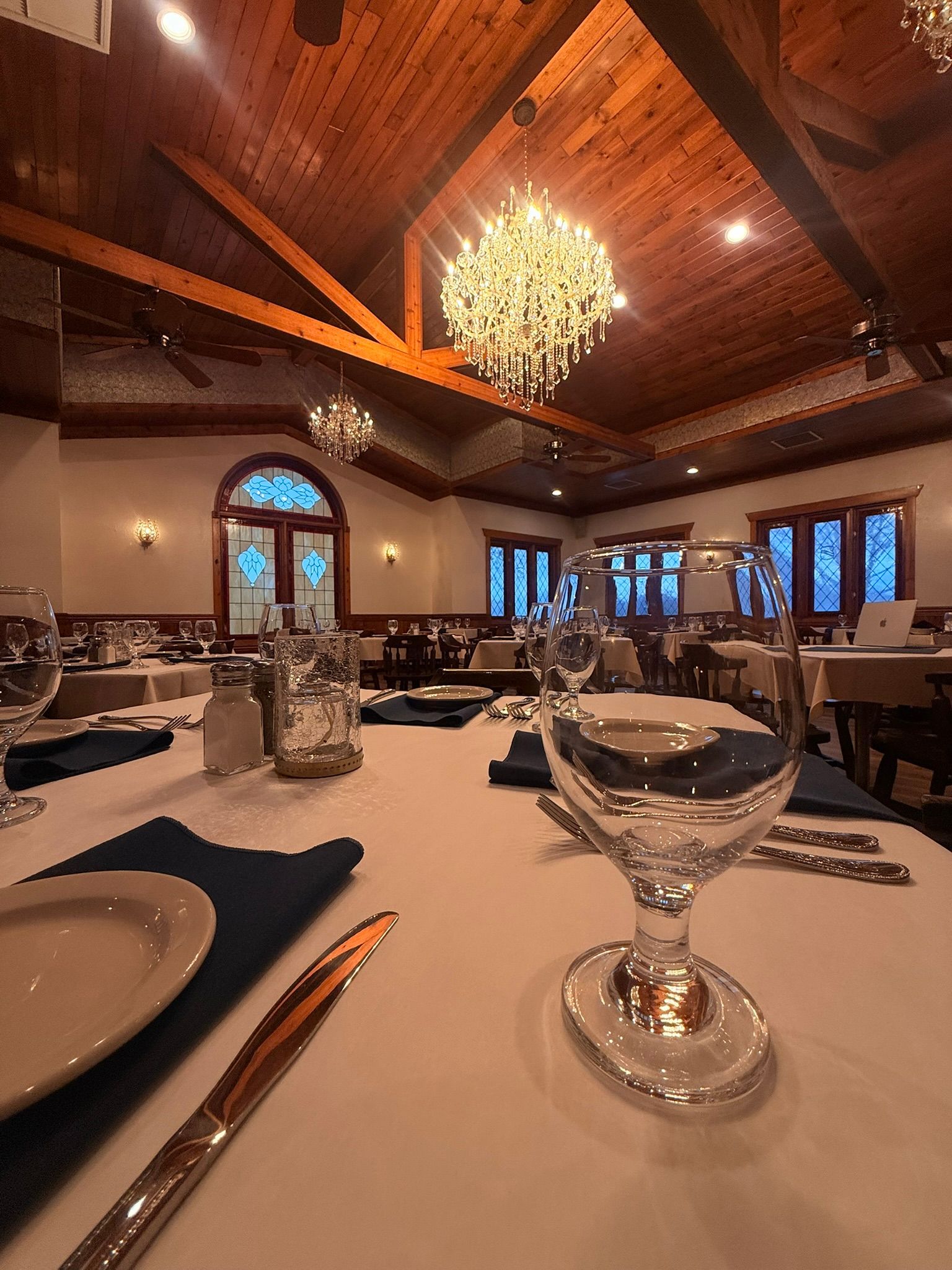 Elegant restaurant interior with chandeliers, tables set with white linens, and dark wood ceiling.