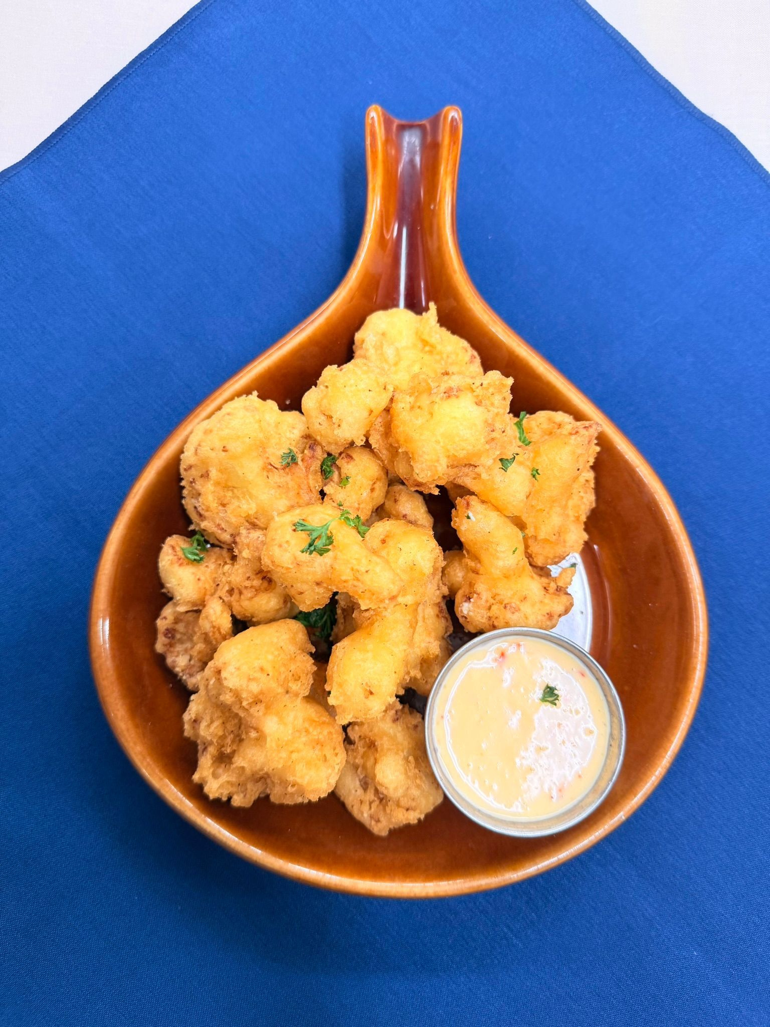 Fried cauliflower florets with dipping sauce in a brown serving dish on a blue tablecloth.