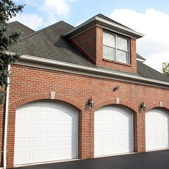 A brick house with three white garage doors and a window on the roof.