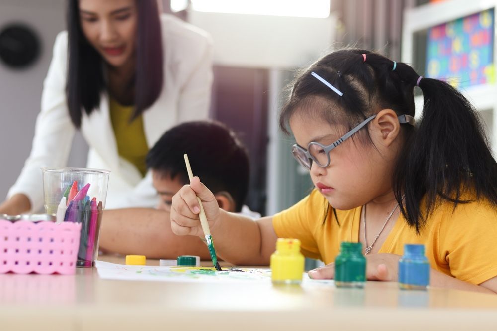 A Girl With Down Syndrome is Painting With a Brush in a Classroom — Better Access Support Services In Dalby, QLD
