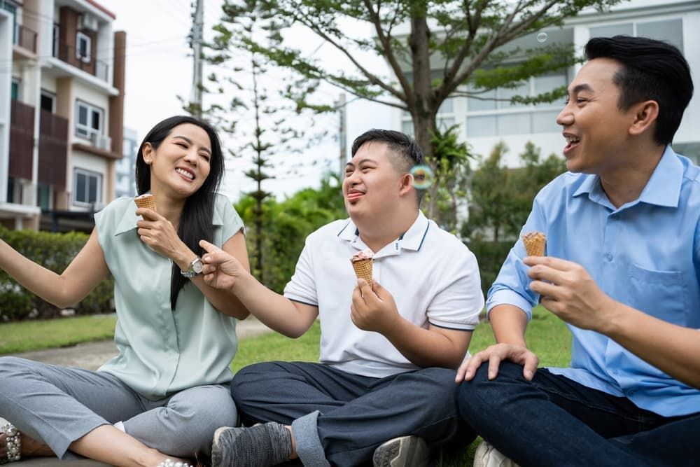 Three people sit on grass outdoors, laughing and eating ice cream cones near modern residential buildings.