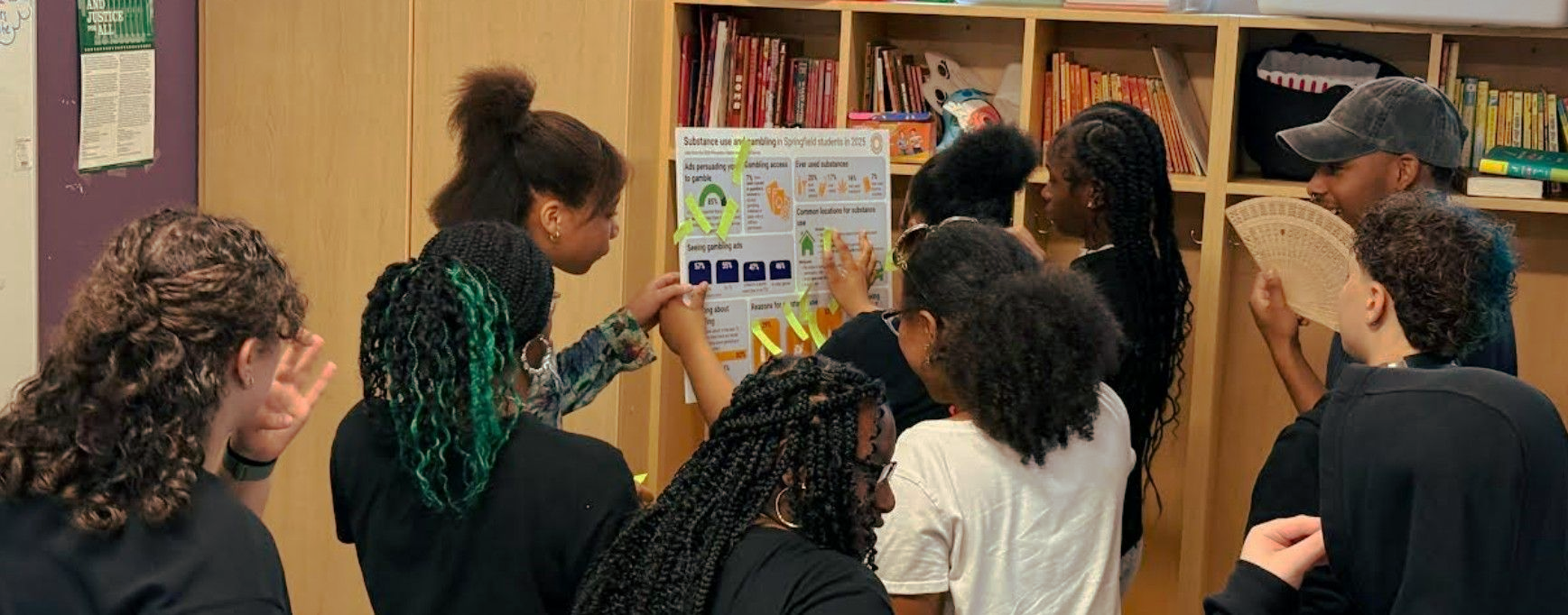 People gathered in a library, facing a poster board and discussing displayed materials.