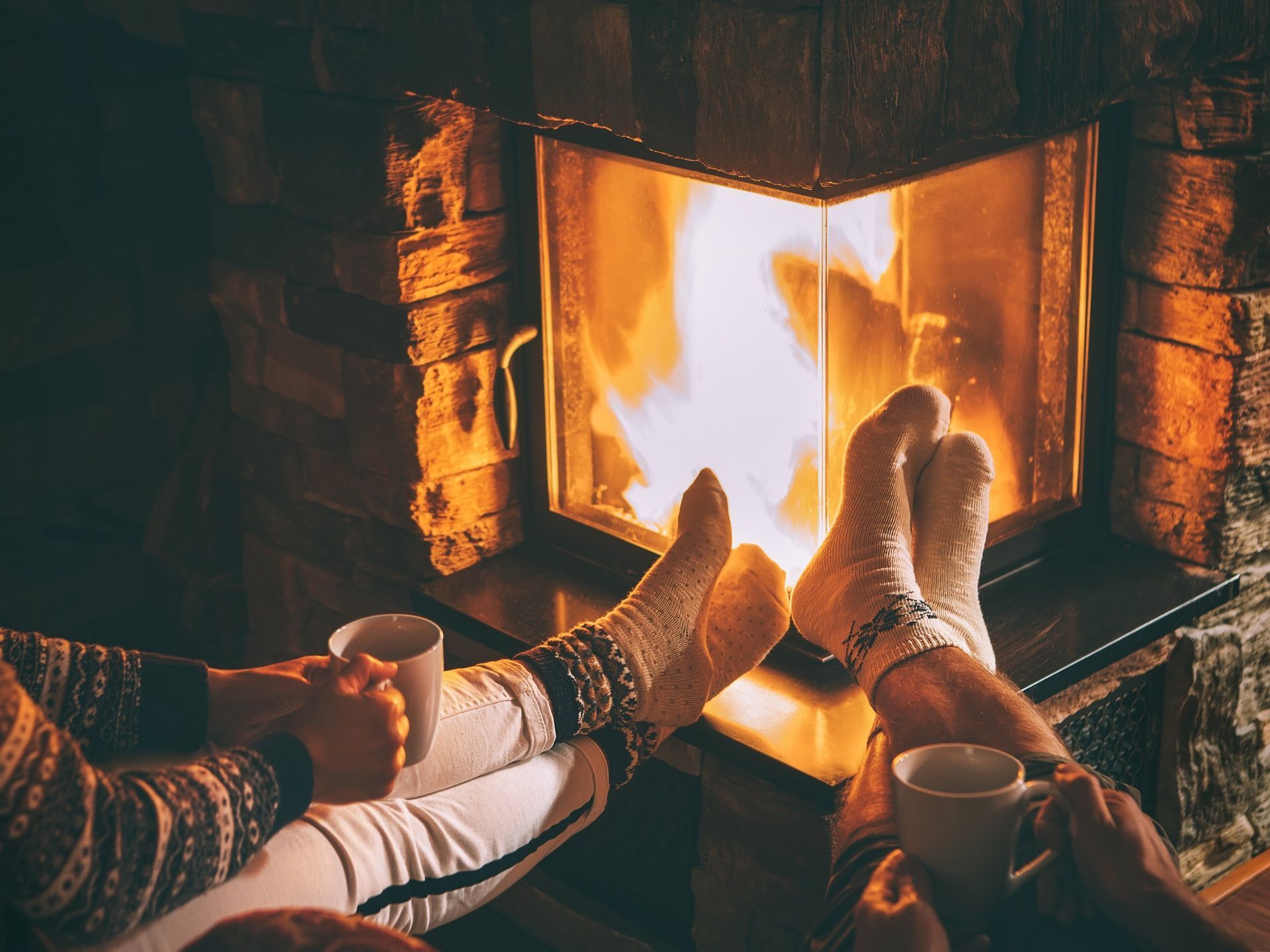 Una coppia è seduta davanti a un caminetto con i piedi alzati e beve un caffè.