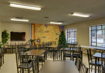 Empty dining hall with tables and chairs, wall art, and large windows letting in daylight