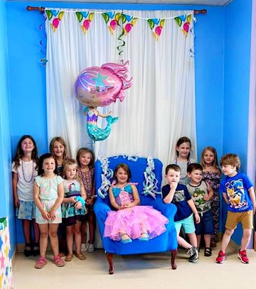 Children posing in a blue party room with balloons and a girl in a pink dress seated on a chair