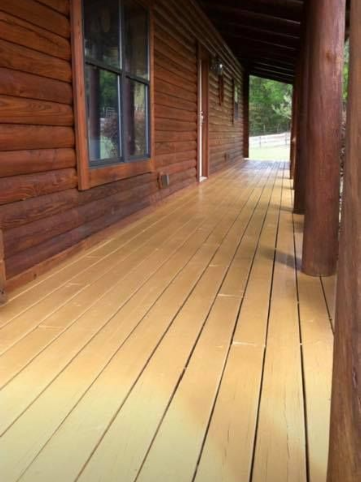 Wooden porch of a log cabin with new light-colored flooring, leading toward a wooded area.