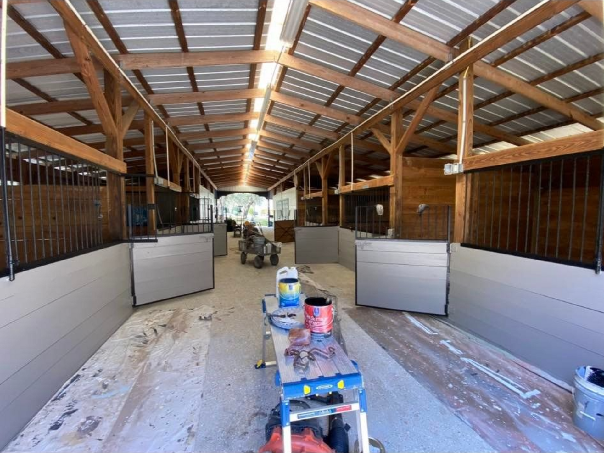 Interior view of a barn under construction with gray stalls and wooden beams. Tools and paint buckets are visible.