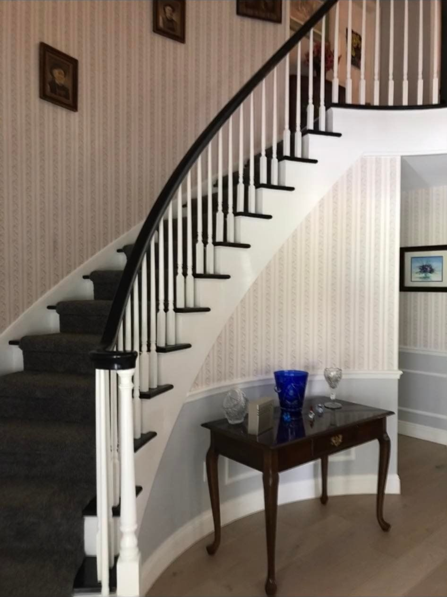 Curved staircase with black railing, white balusters, and gray carpet. Table with vase and glasses.