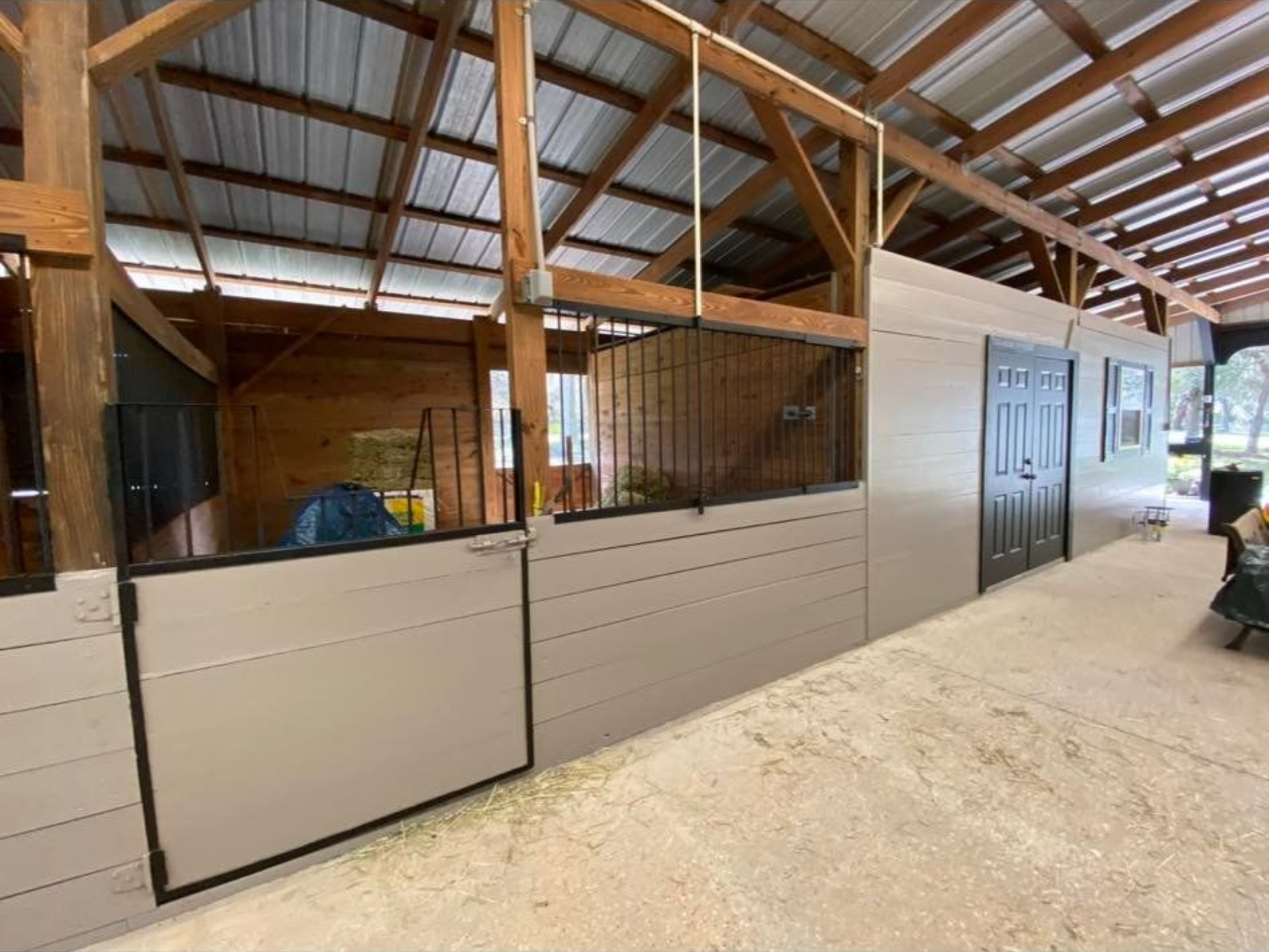 Interior of a barn with horse stalls. Beige stalls with black doors and grates, set in a wood frame under a vaulted ceiling.