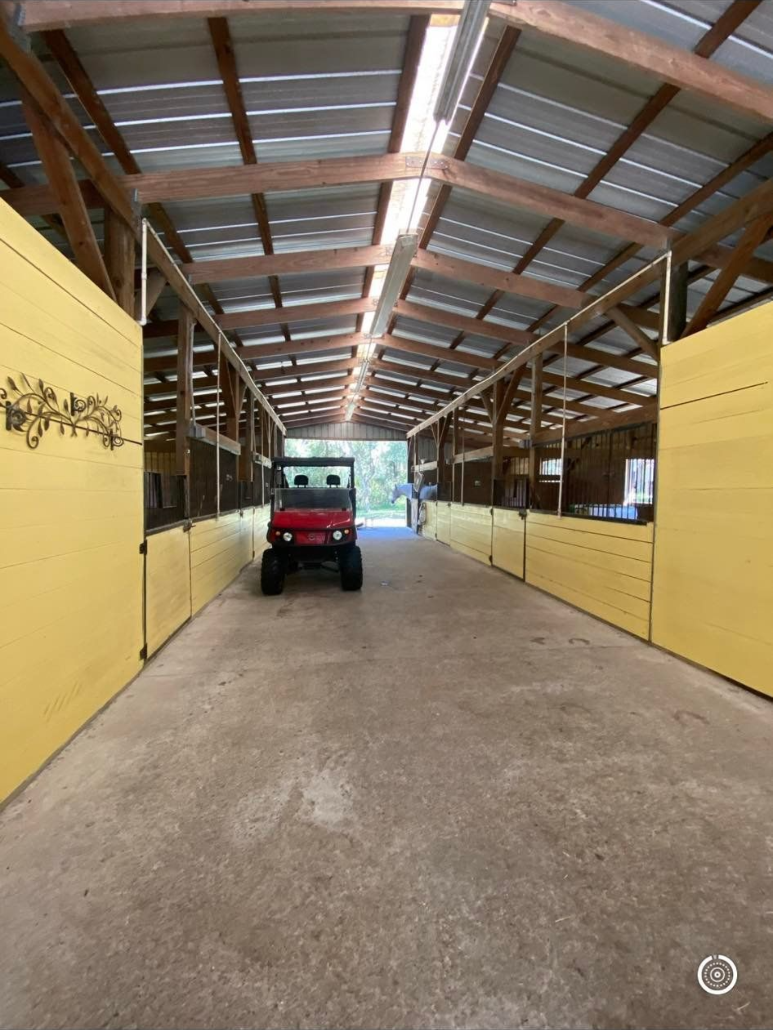 Red utility vehicle in a barn aisle, flanked by yellow stalls under a wood and metal roof.