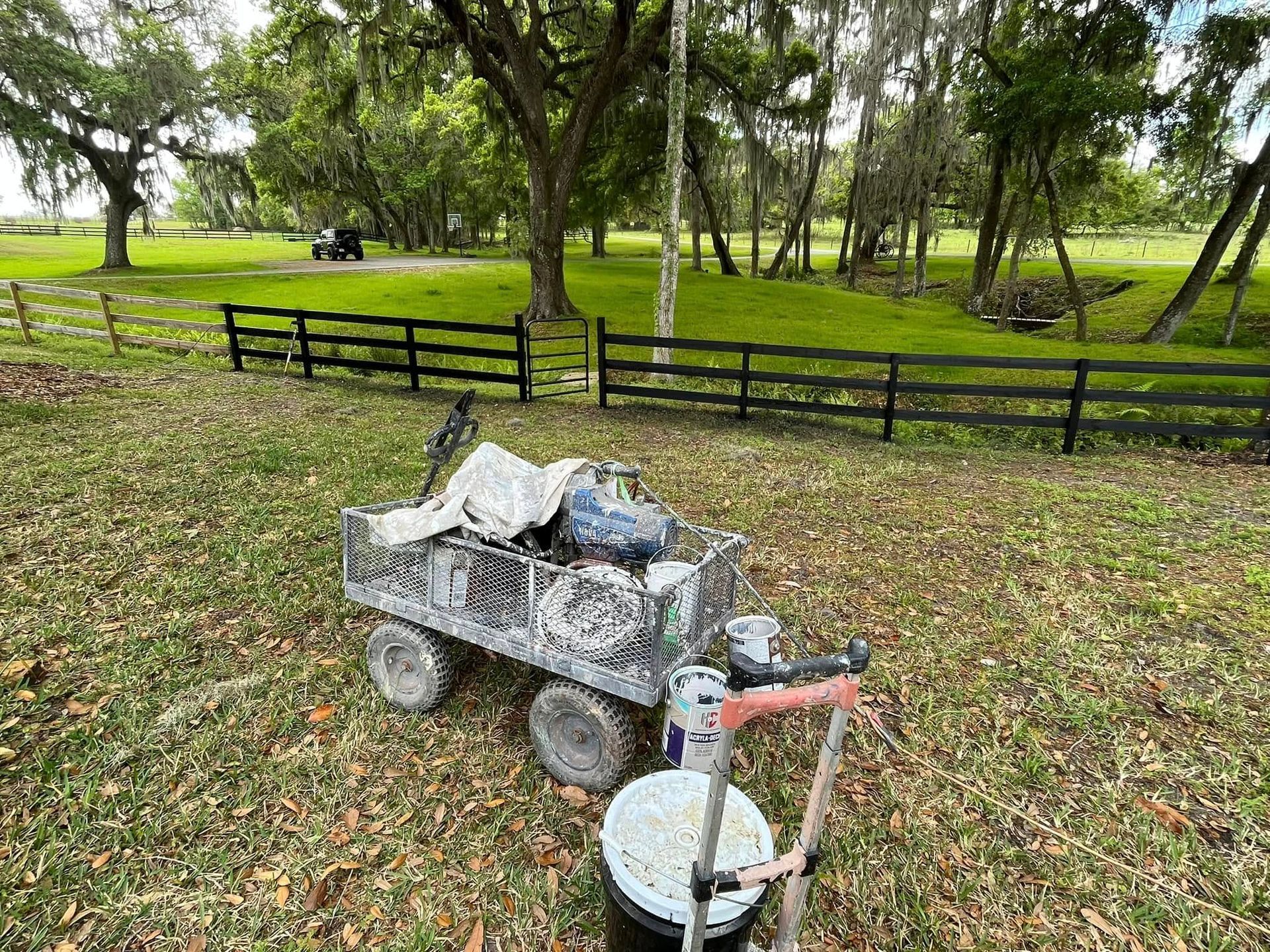 A paint cart and sprayer sit in a grassy yard, with a newly painted black fence in the background.