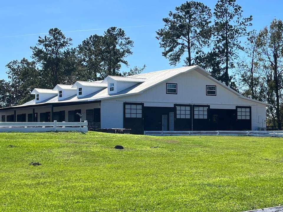 White horse barn with black doors and windows, green field, and blue sky.