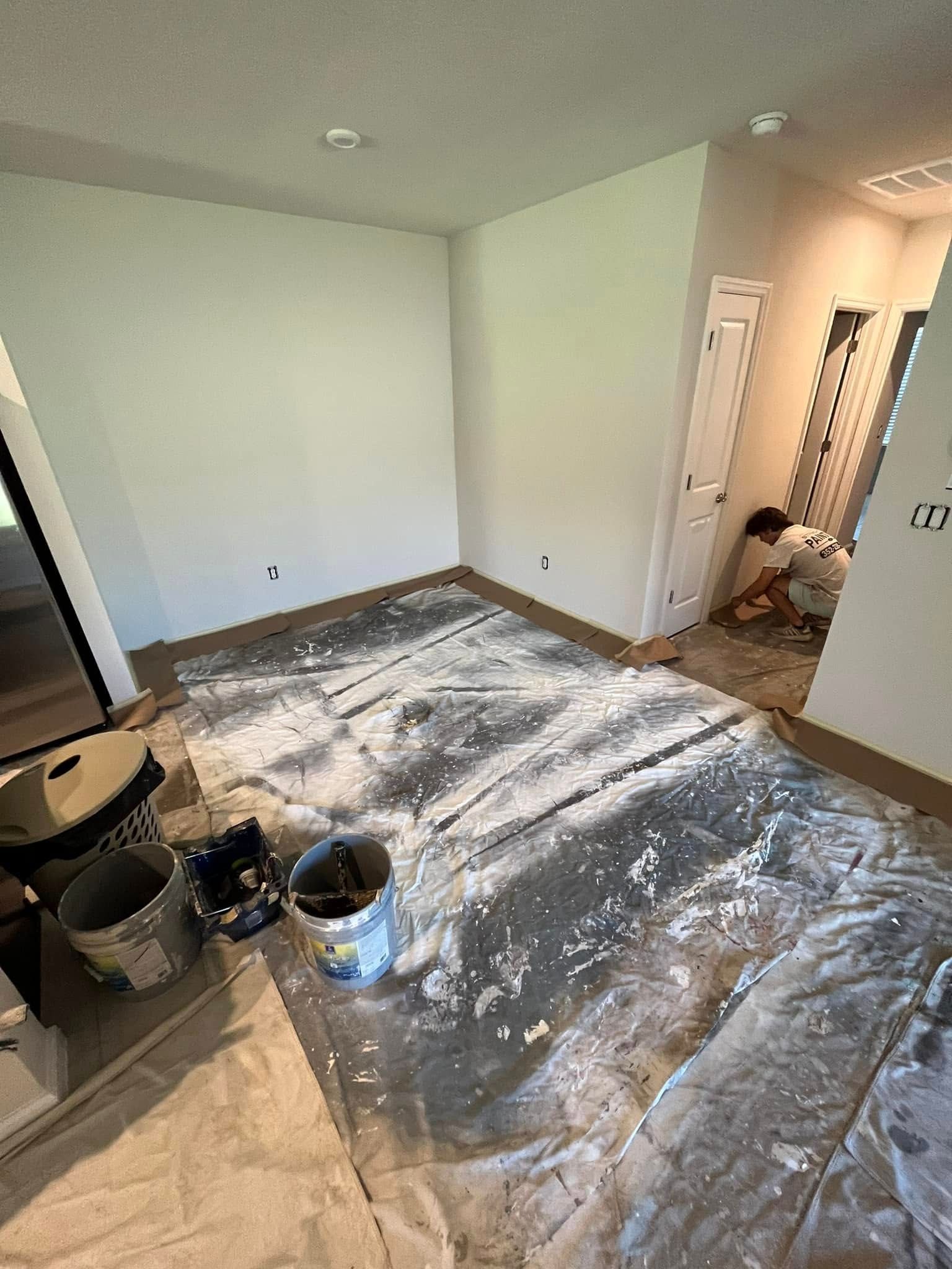 Room being painted; painter on knees near doorway. Paint cans, drop cloths, and tools on floor.