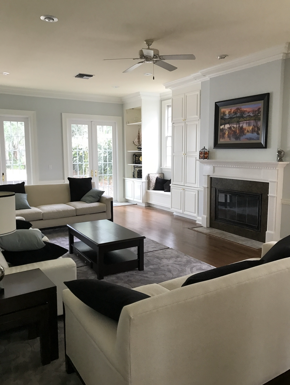 Living room with white sofas, fireplace, and French doors. Light blue walls, hardwood floors, and a dark coffee table.