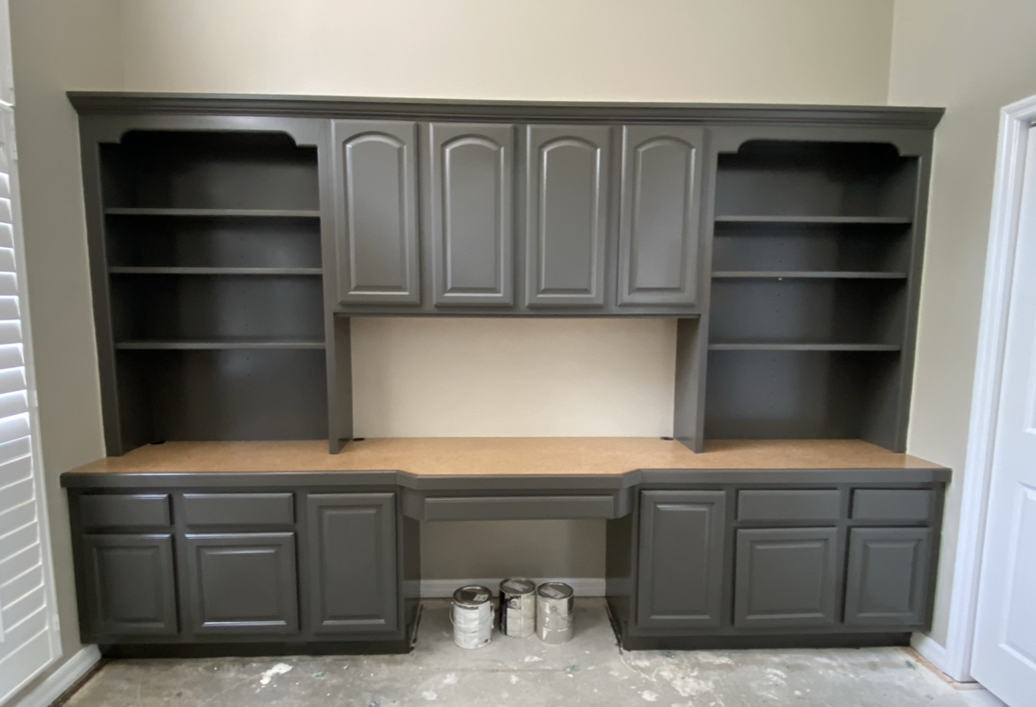 Built-in gray cabinets and shelves above a desk, against a beige wall, in a room under construction.