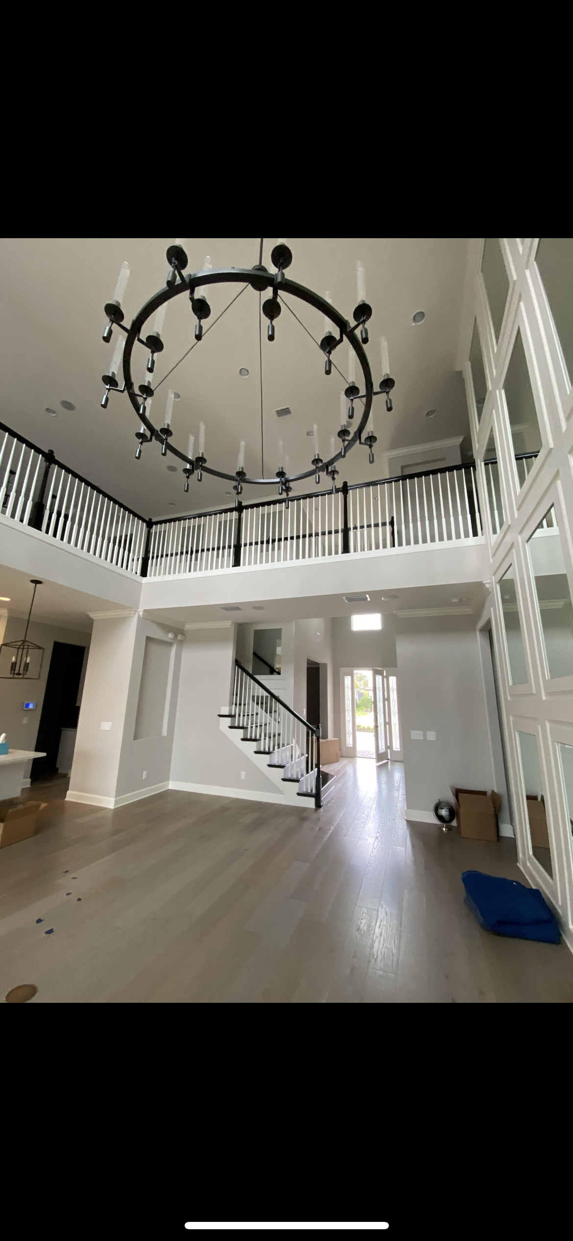 A grand foyer with a large circular chandelier, staircase, and balcony. White walls and wooden floor.