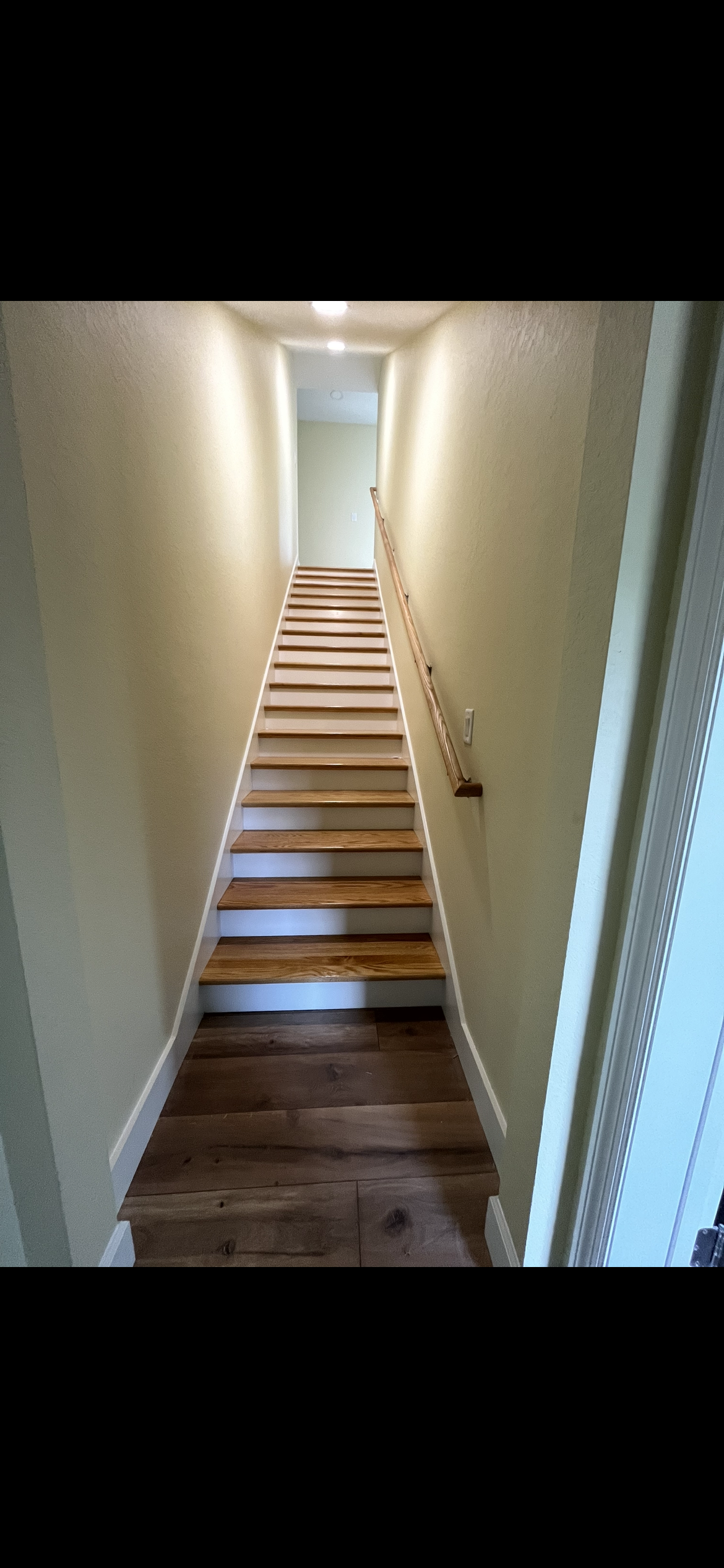 A narrow staircase leads upwards between cream-colored walls. A wooden handrail is on the right.