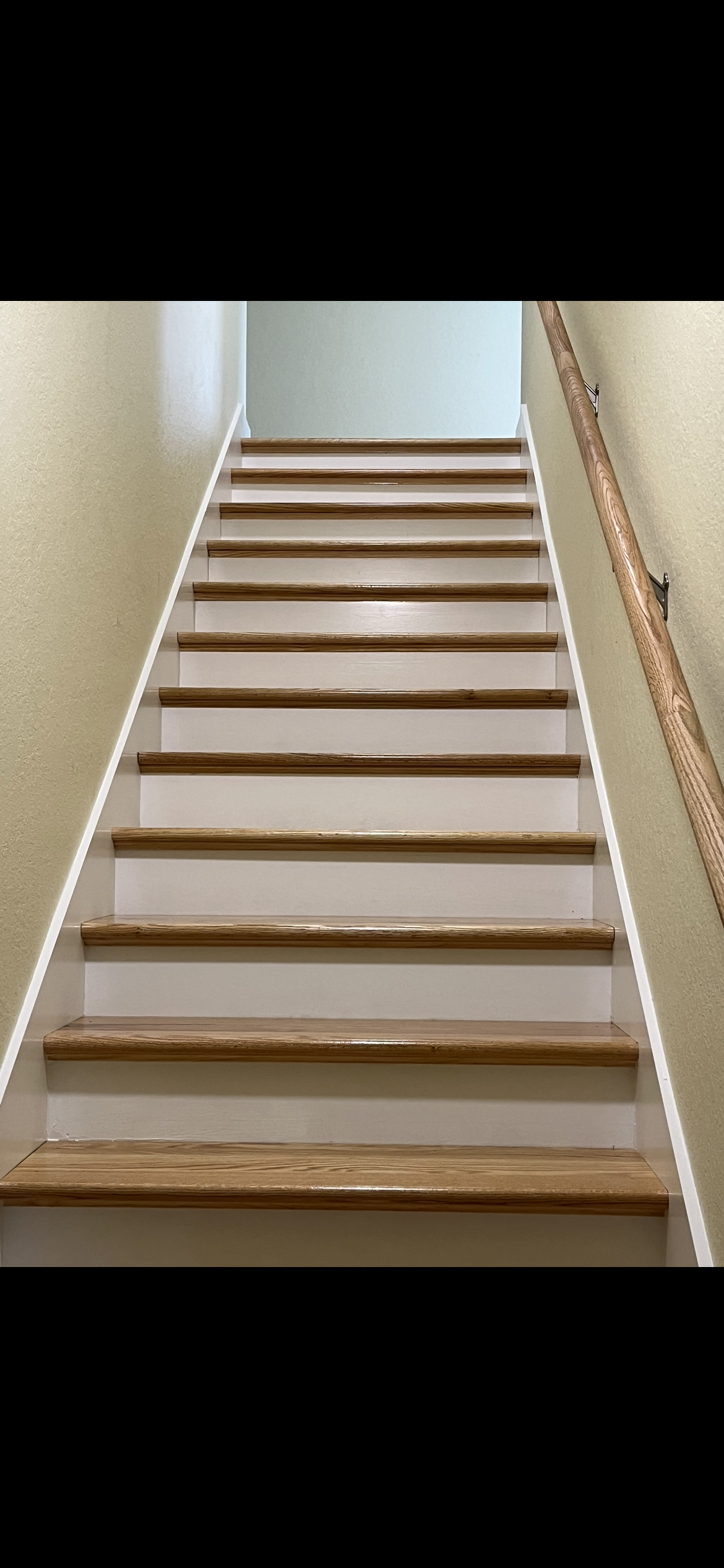 Wooden stairs lead upwards, with white risers and a brown handrail. Beige walls are on either side.
