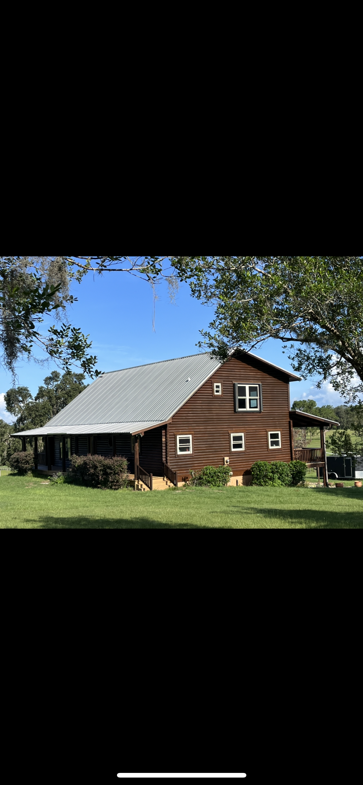 Brown two-story wooden house with a metal roof surrounded by green grass and trees under a blue sky.