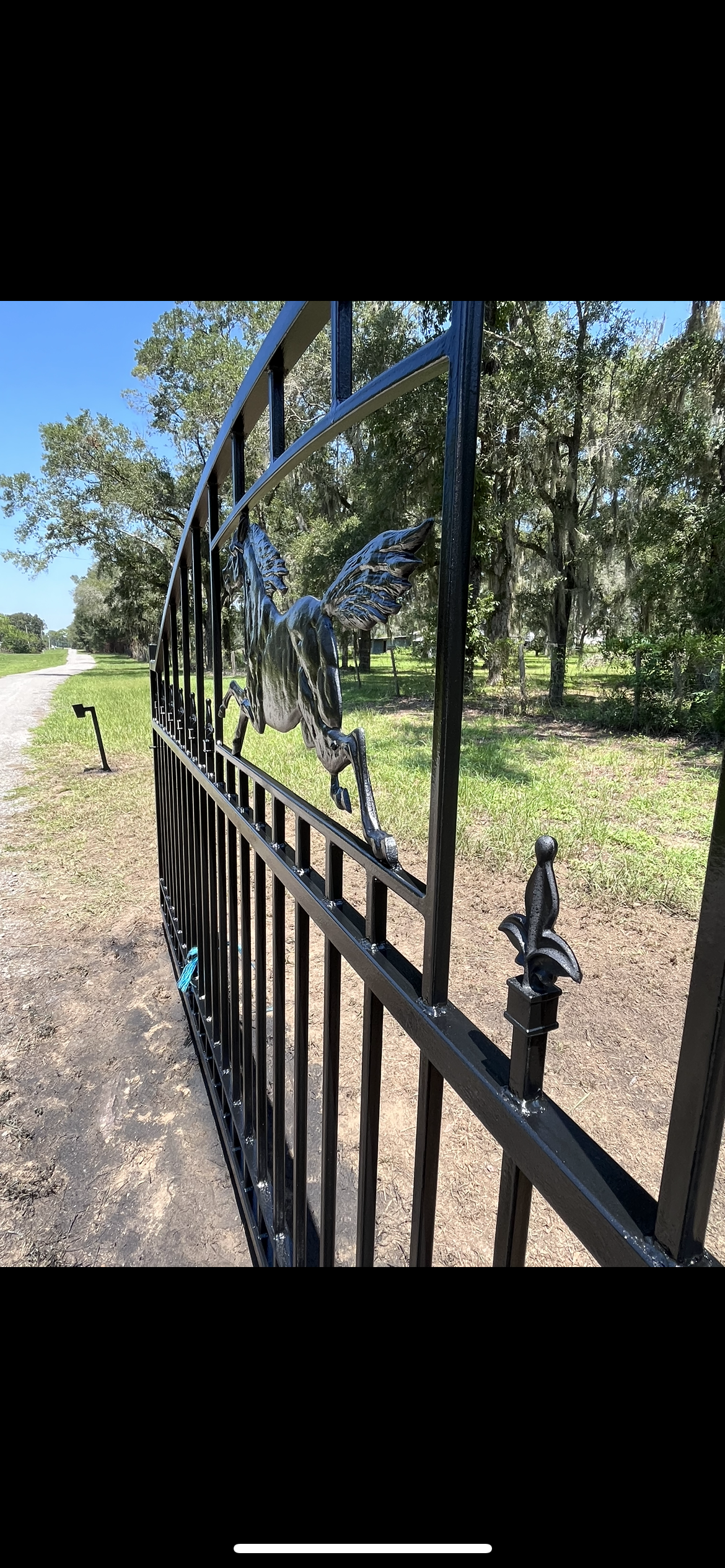 Black metal gate with decorative design, open on a sunny day, leading to a path and trees.