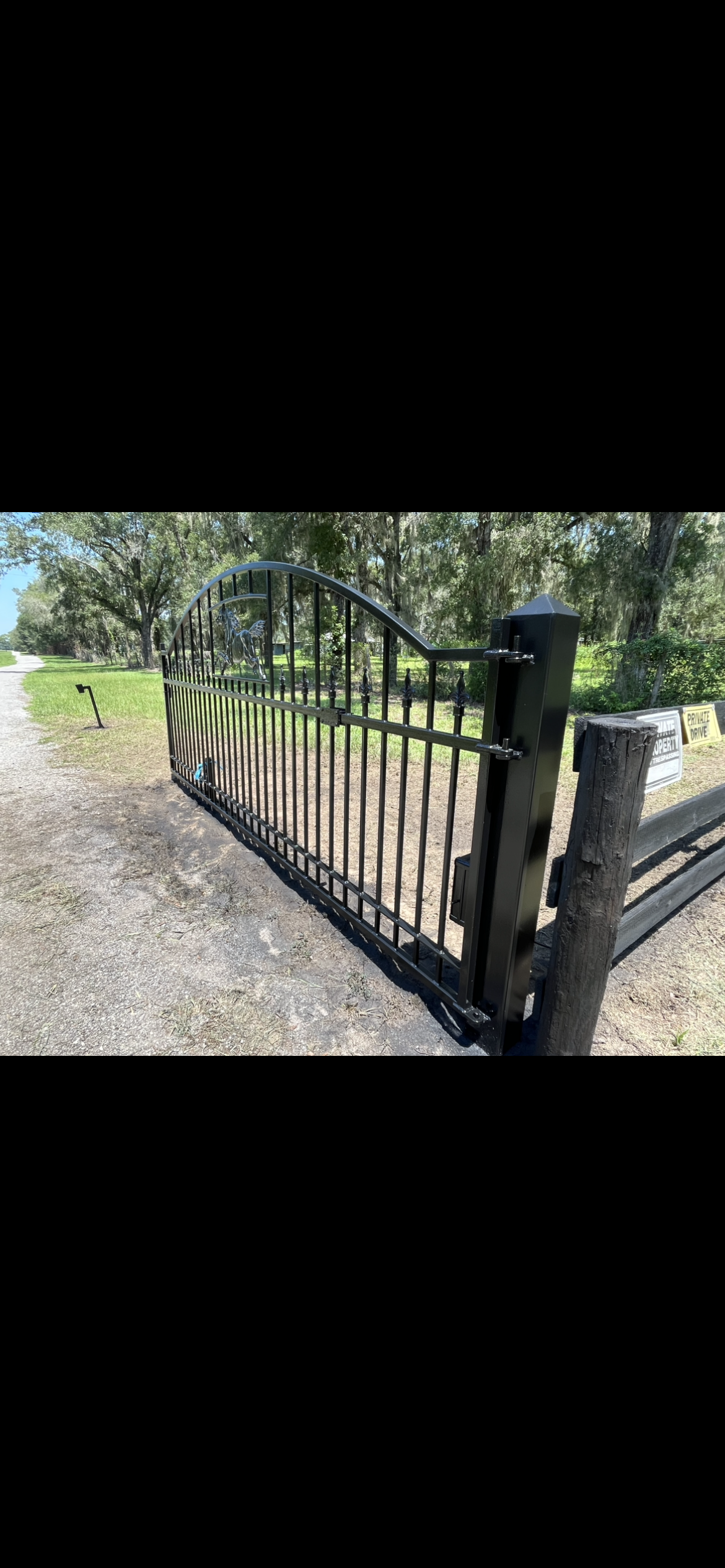 Black metal gate and fence in front of a gravel road and trees.
