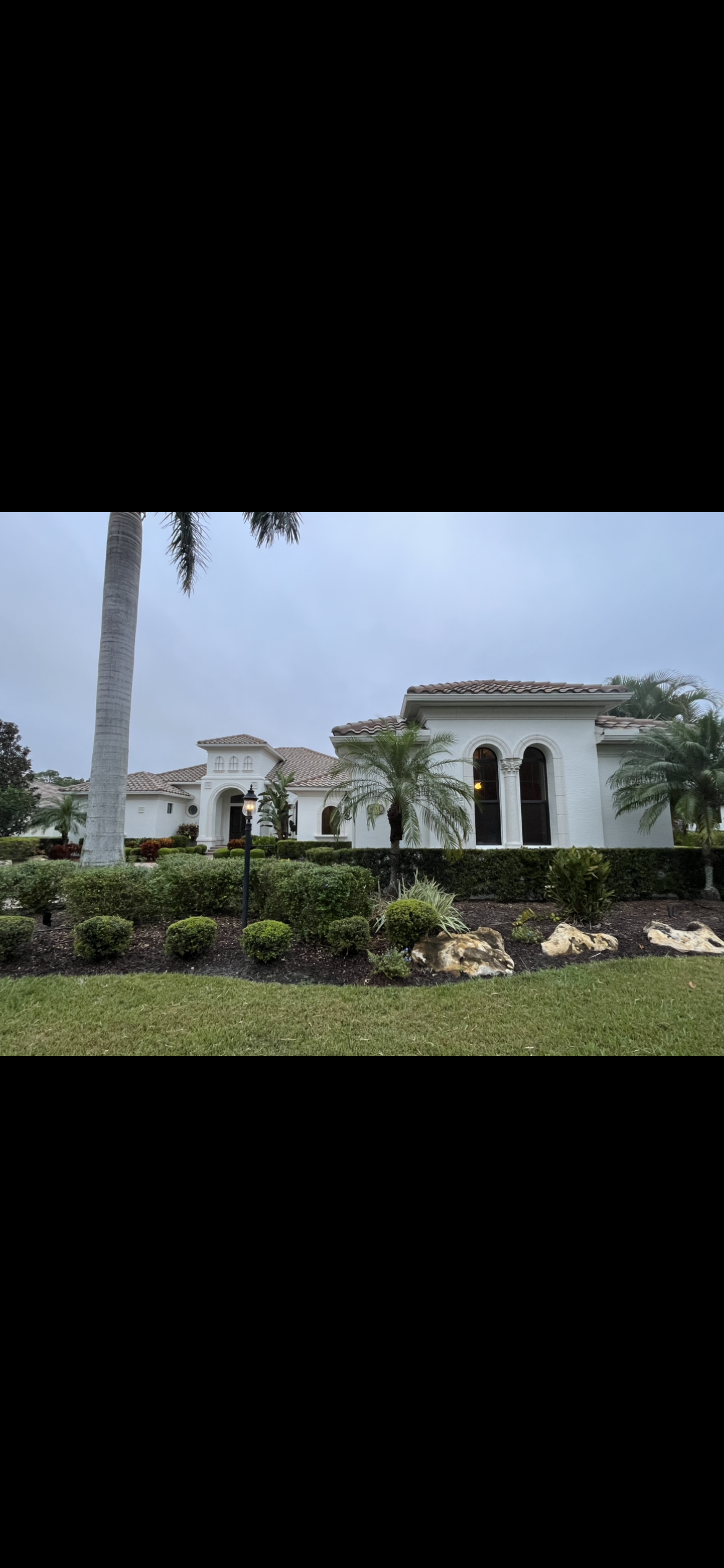 White stucco house with arched windows, green hedges and lawn under a cloudy sky.