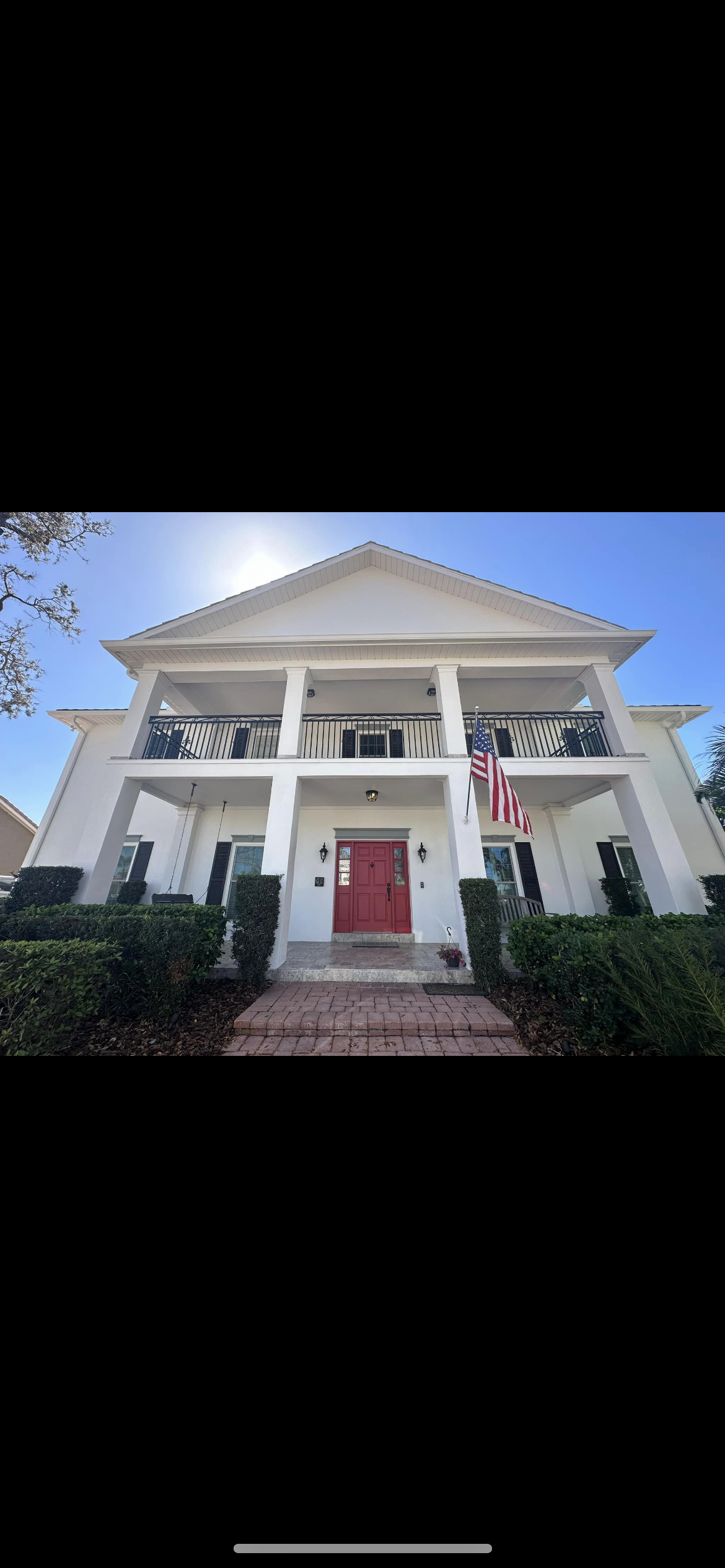 White two-story house with red front door, porch, and American flag hanging. Bushes line the front.