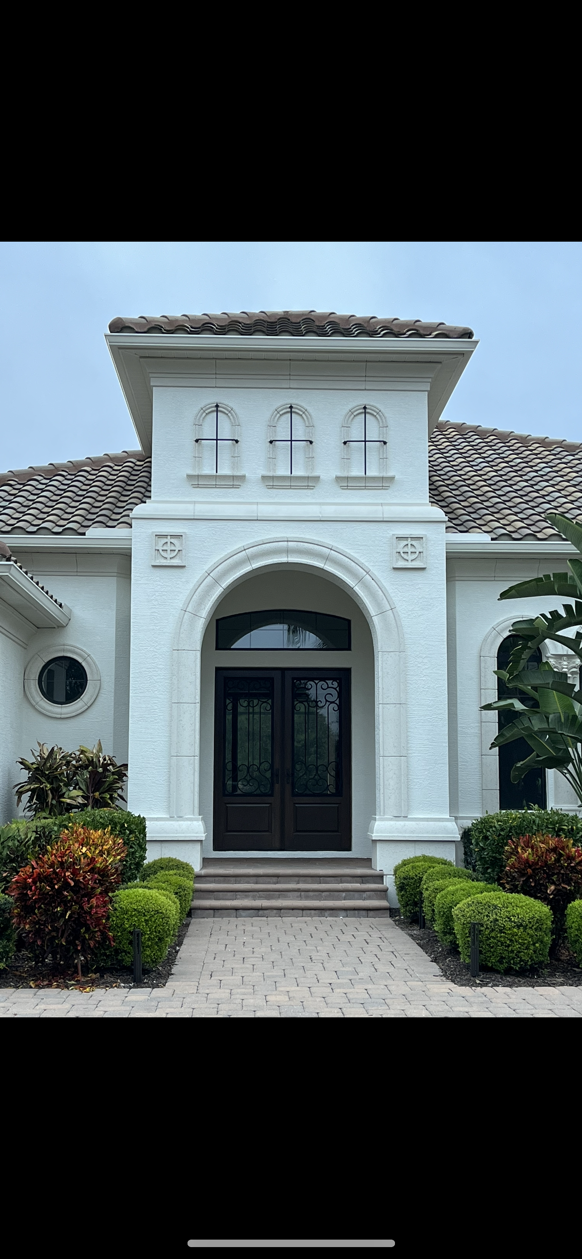 White stucco house with arched doorway and black double doors, flanked by landscaping.