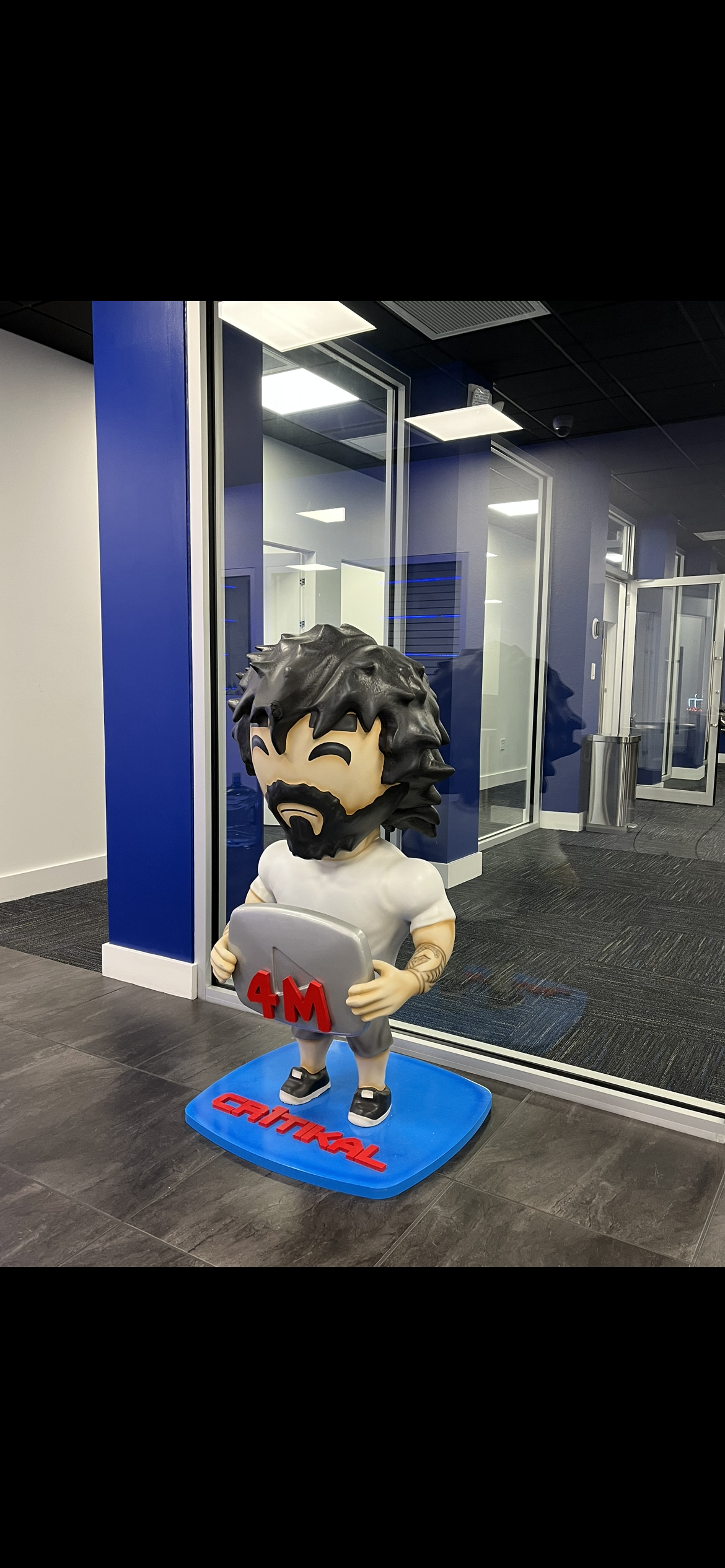A figure holding a tablet stands on a blue base. A blue wall and glass windows are in the background.