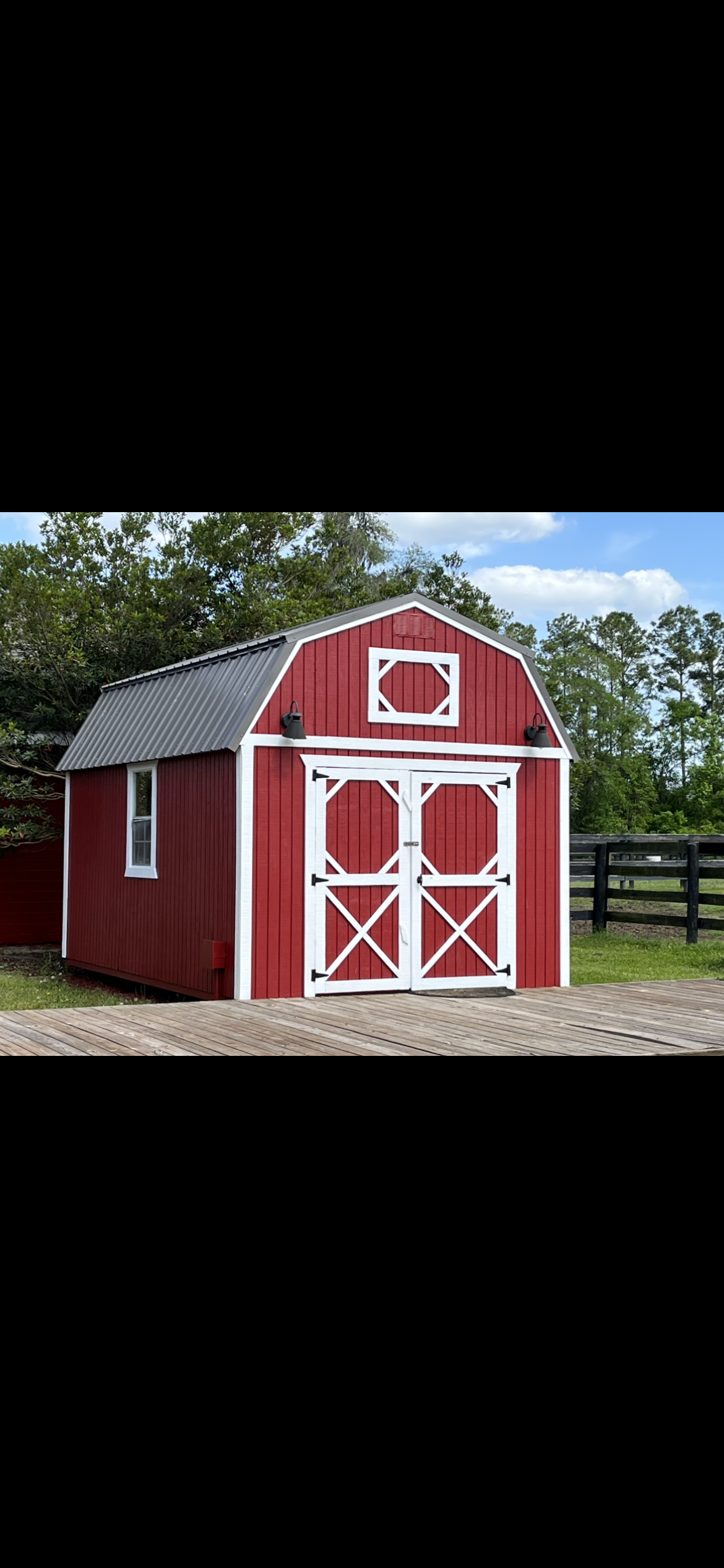 Red barn with white trim and gray roof, in a grassy area with trees and a fence in the background.