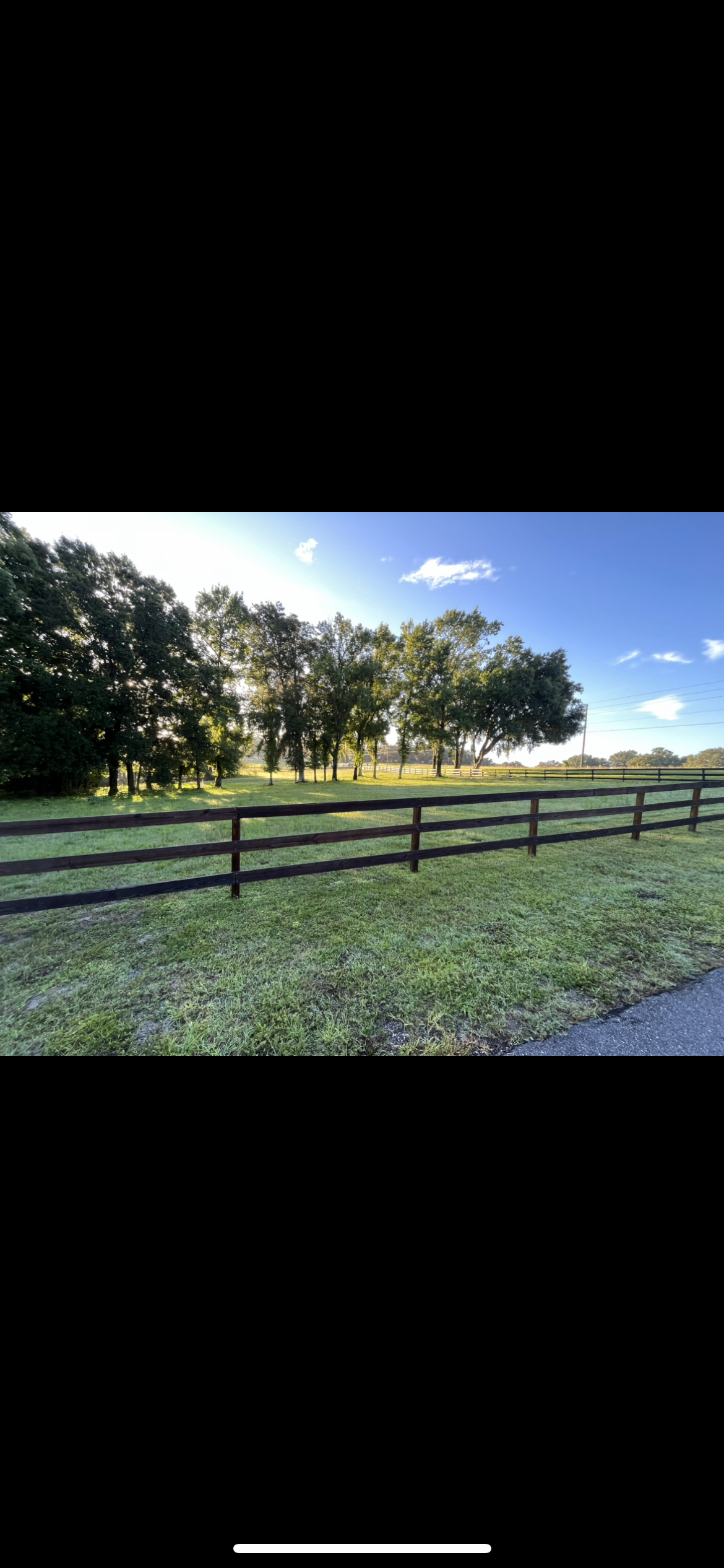 A rural scene with a wooden fence, green grass, trees, and a blue sky with some clouds.