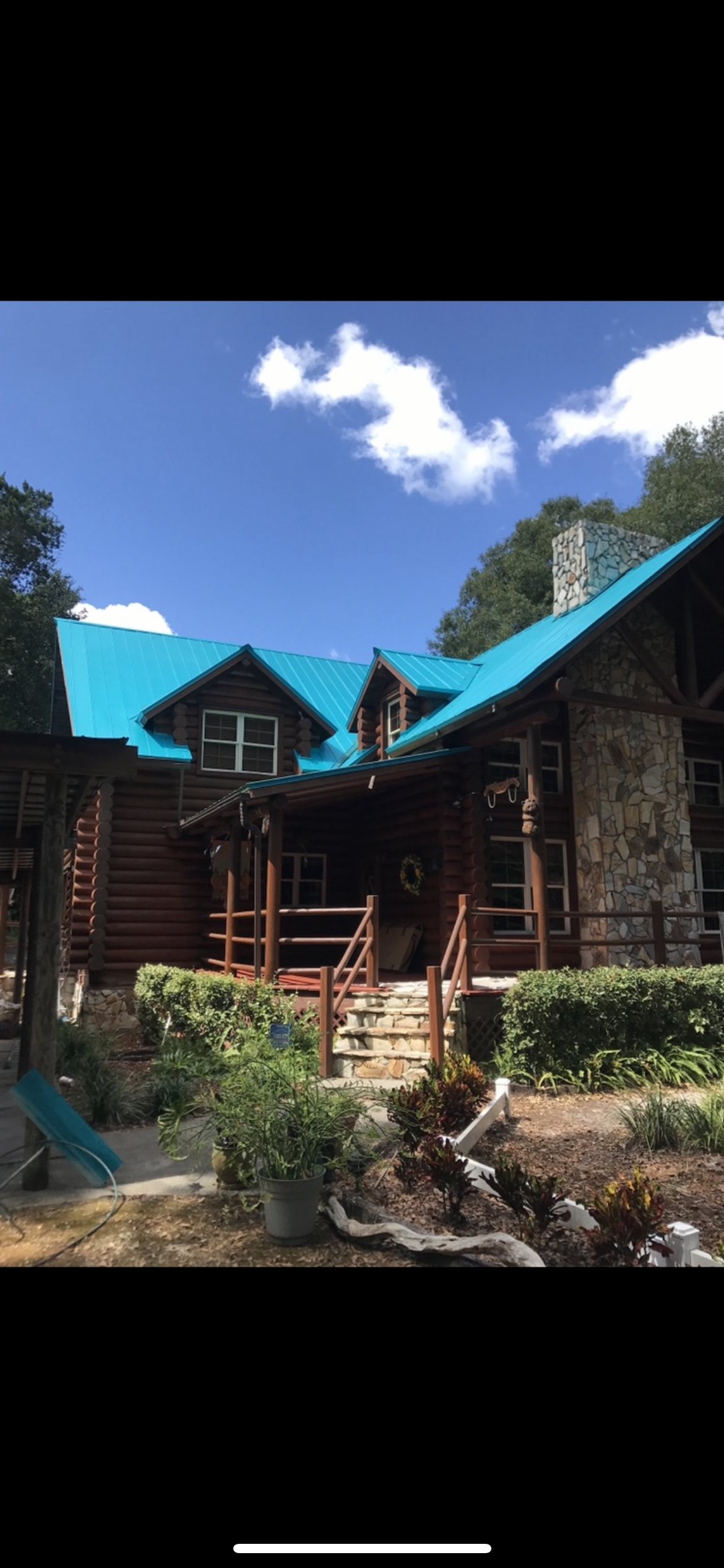 Log cabin with a turquoise roof, stone chimney, and wooden porch with a blue sky in the background.