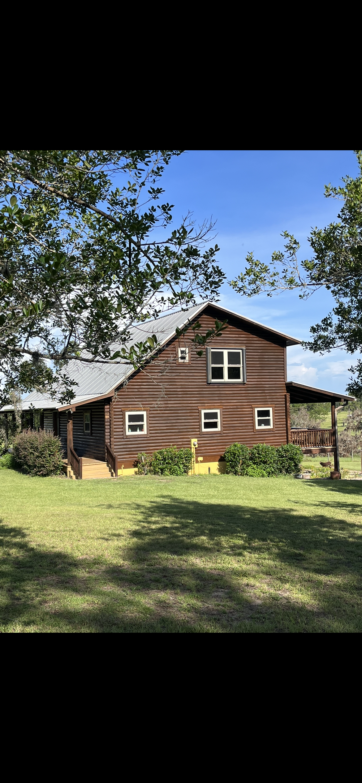 Rustic, two-story wooden cabin with a porch and green lawn, surrounded by trees under a blue sky.
