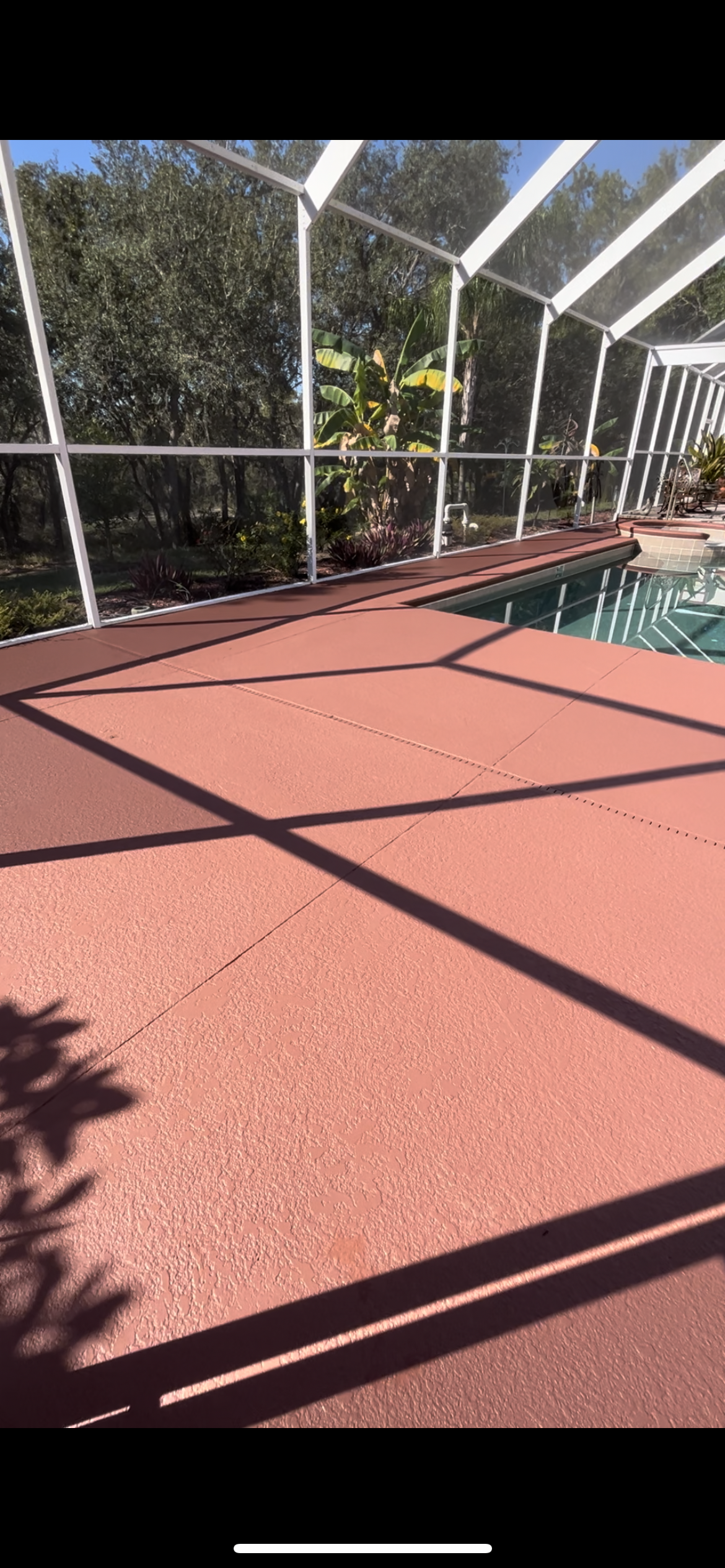 A red concrete patio surrounds a swimming pool, enclosed by a screen. Sunlight casts shadows on the patio.