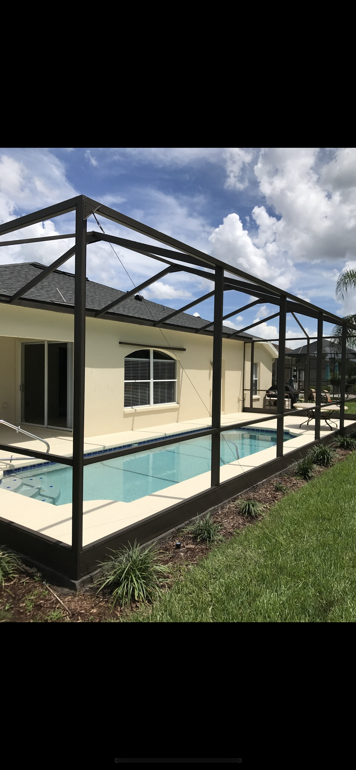 A pool enclosed by a dark framed screen, next to a beige house, under a blue sky.
