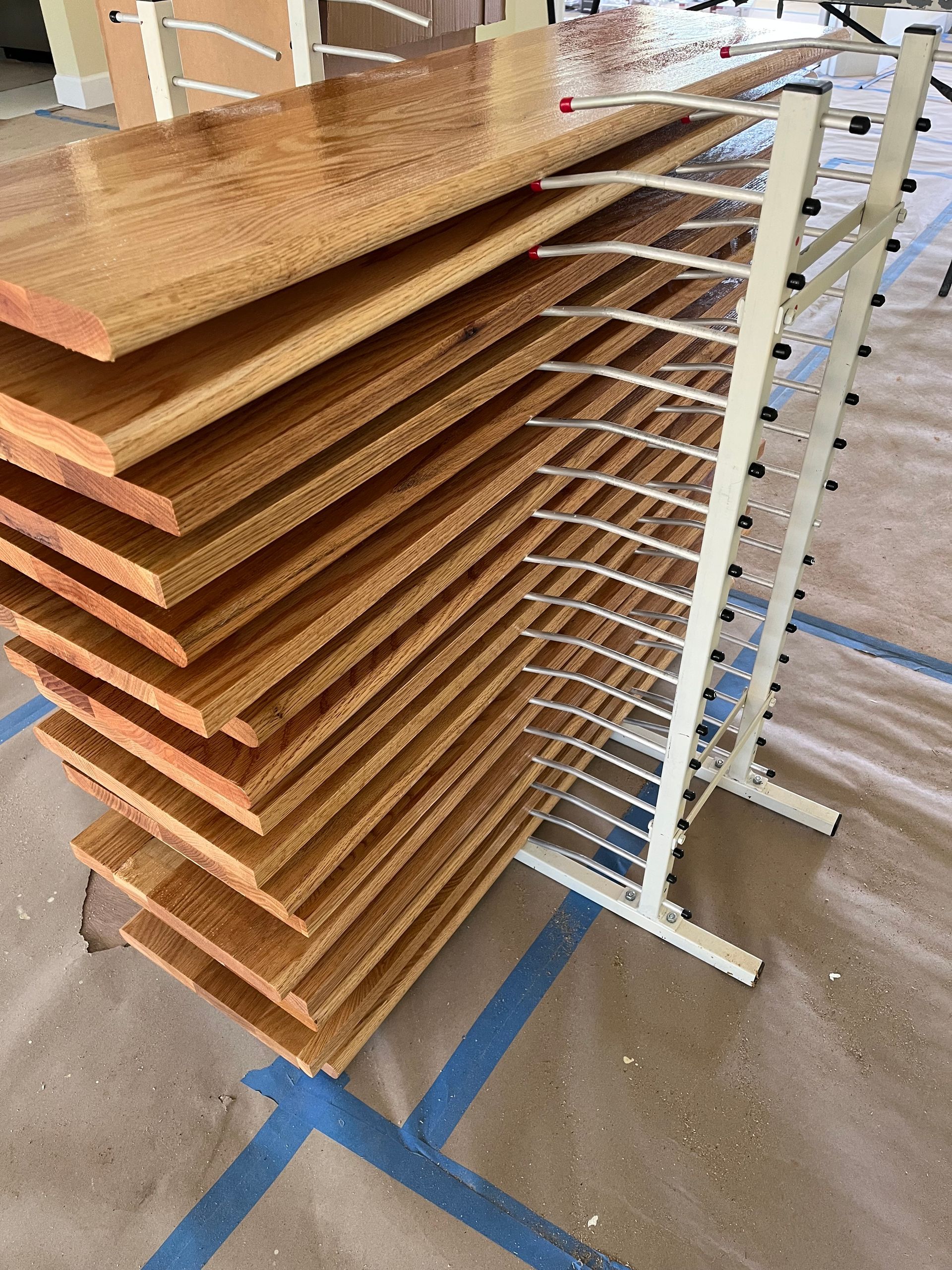 Wooden planks stacked on a drying rack in a room under construction, with blue painter's tape on the floor.