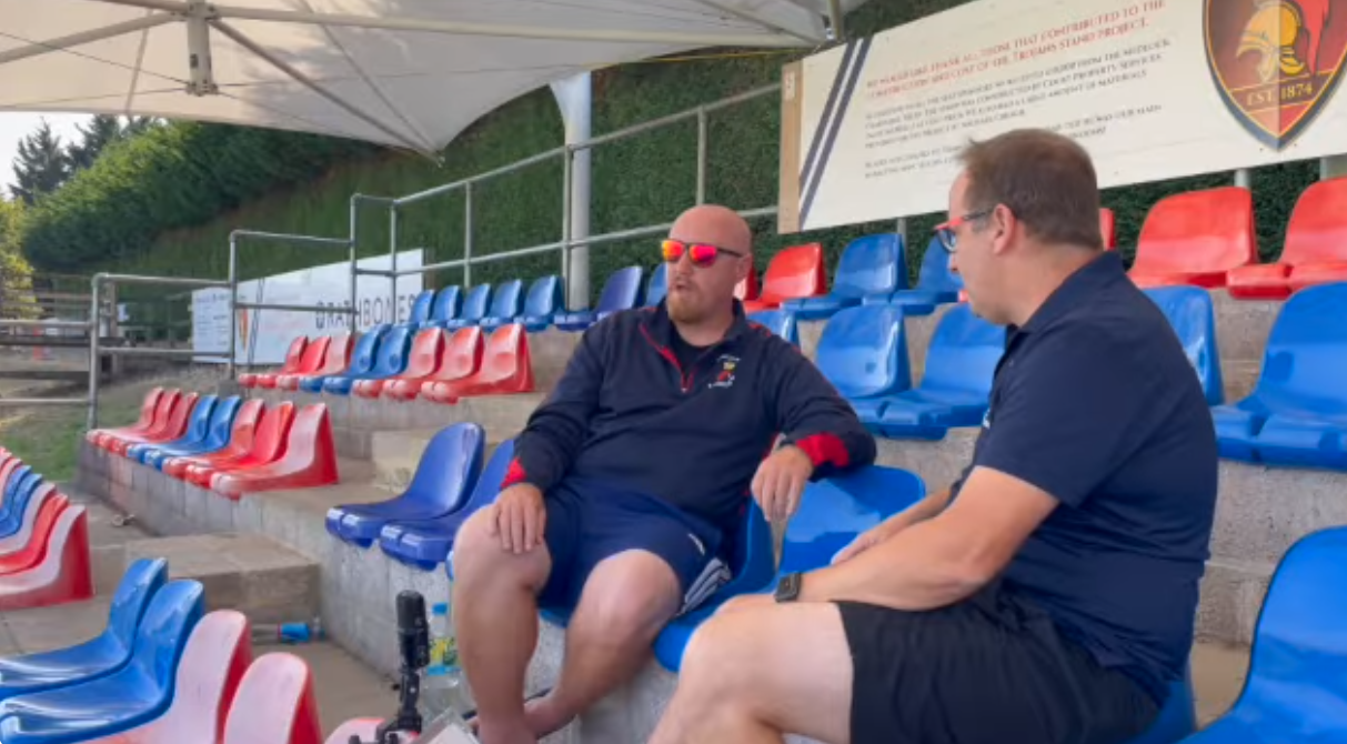 Two men sit on blue and red stadium seats, under a shaded area. One wears red sunglasses.
