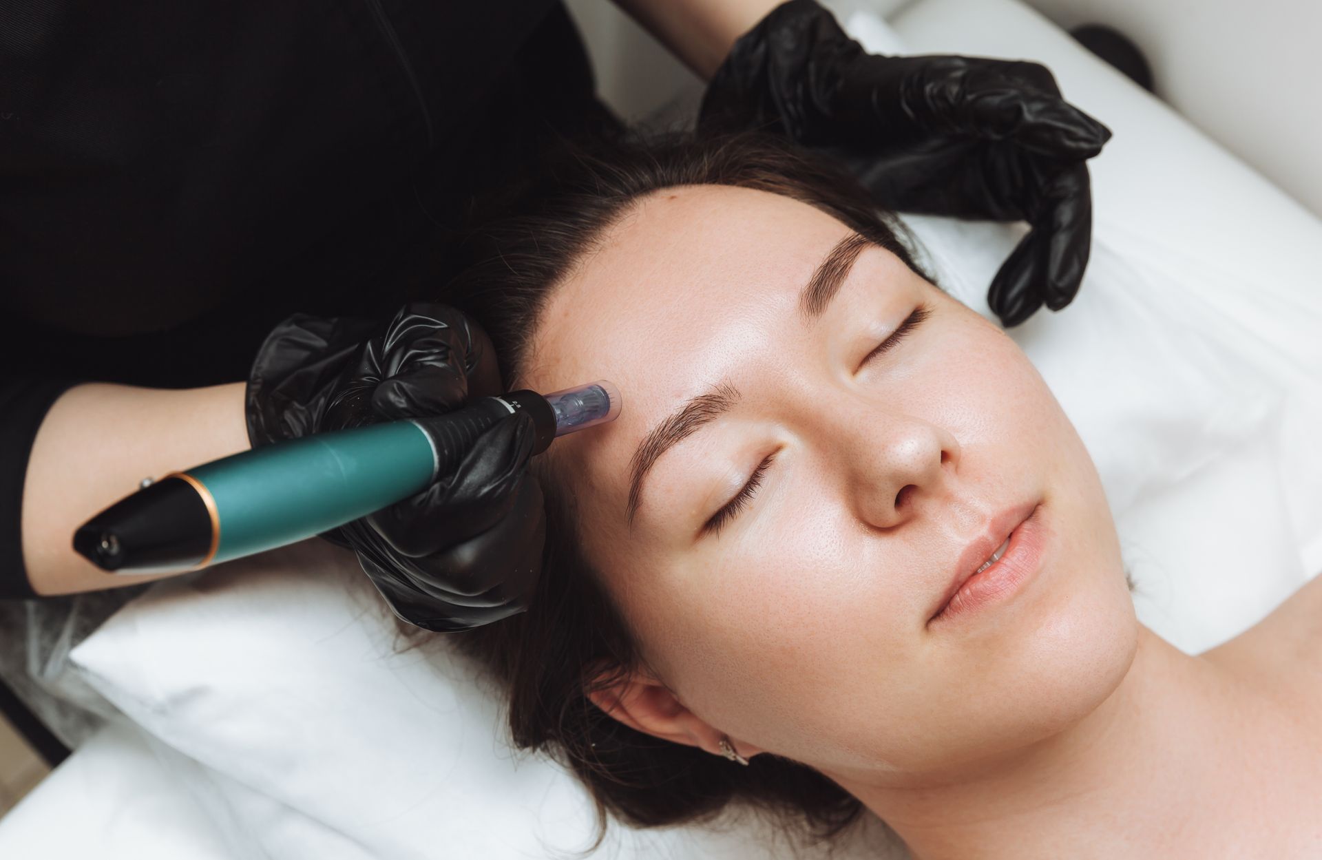 Woman receiving eyebrow tattooing with a teal device while lying on a treatment bed