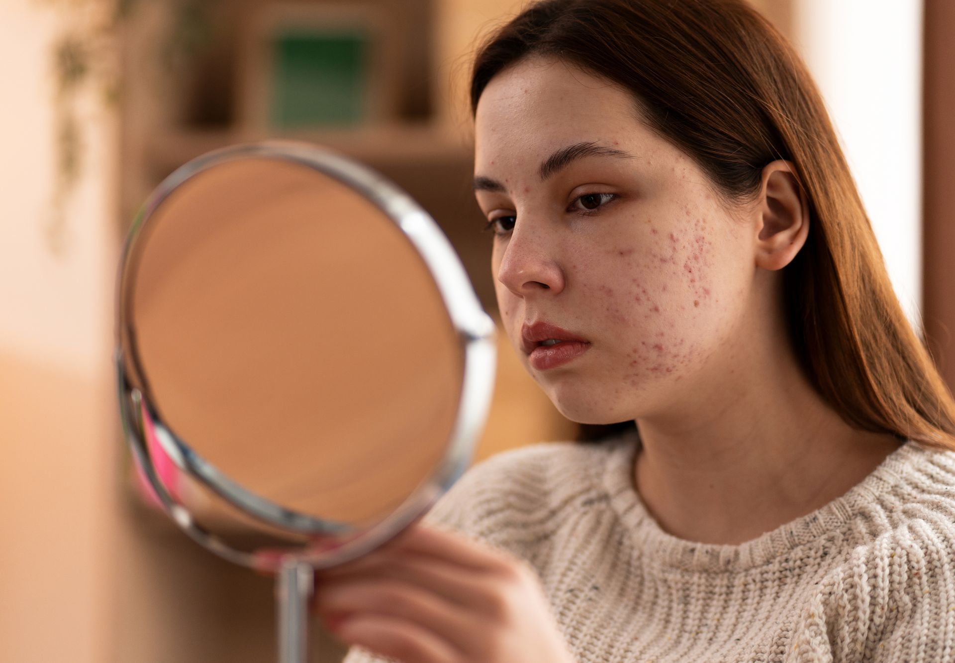 Woman looking at acne in a mirror, indoors, with neutral expression.