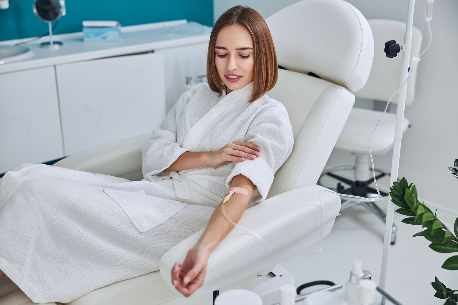 Woman in a white robe receiving an IV drip while seated in a medical chair. Bright, clean setting.