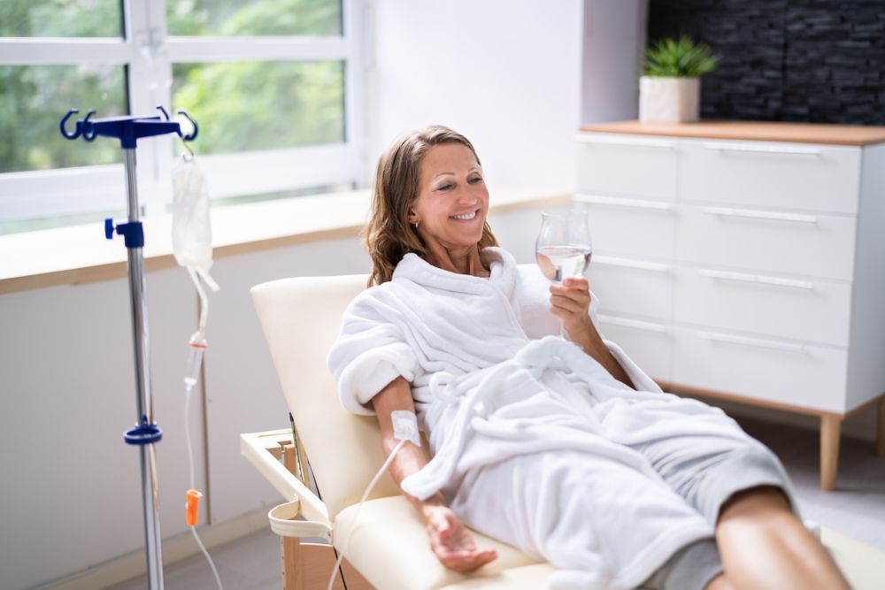 Patient reclining in a home chair with an IV stand nearby, holding a glass of water and smiling