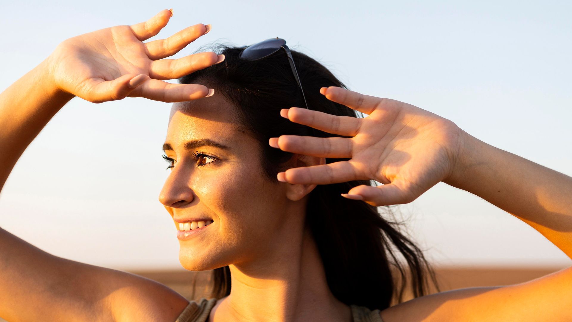 Smiling person shielding their eyes from the sun at a beach, with hands raised against a bright sky