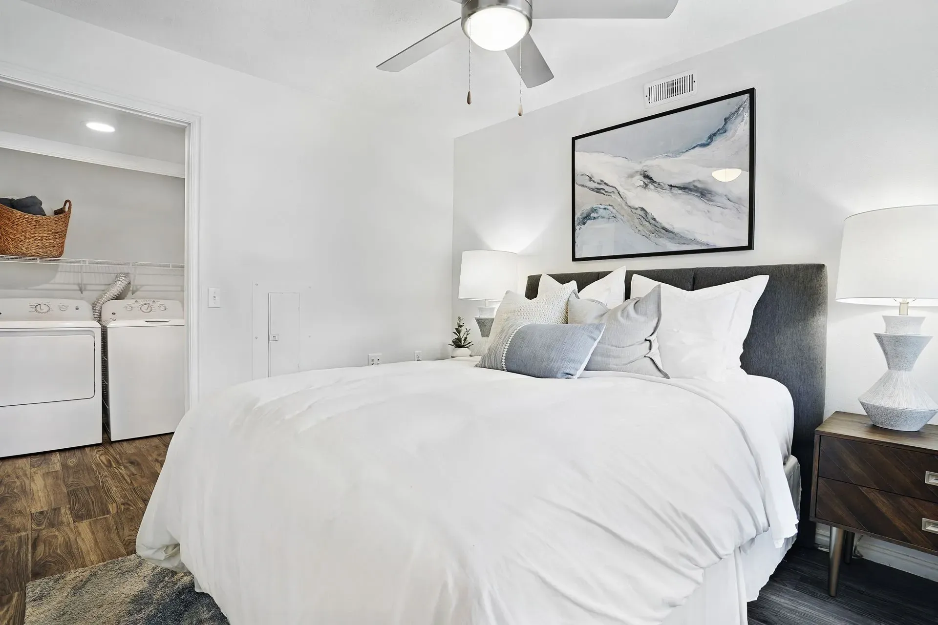 Cozy bedroom with a queen-sized bed, plush white bedding, and decorative pillows. A washer and dryer are visible through an open doorway.